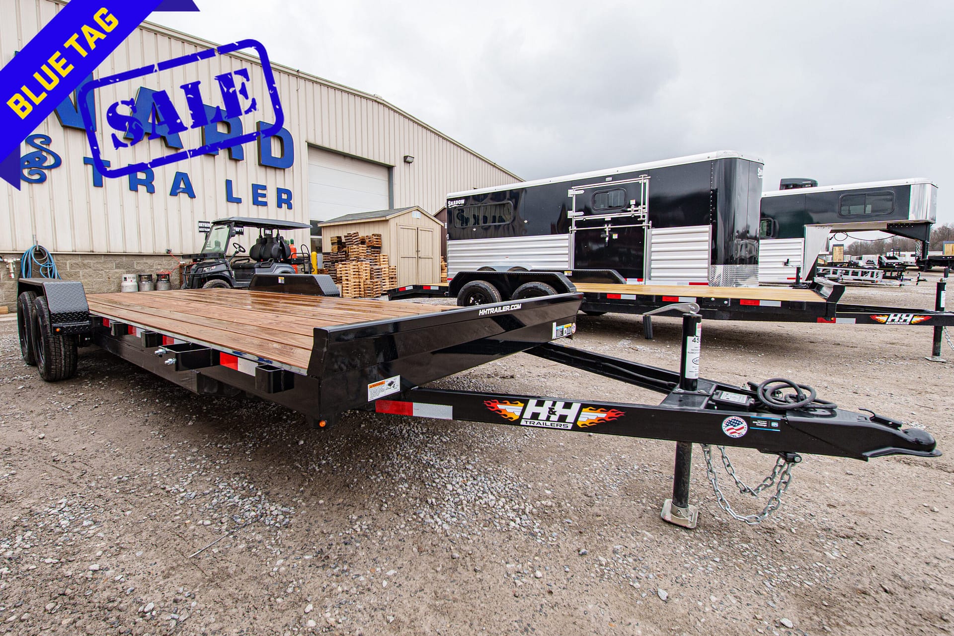 Flatbed trailer in a gravel lot outside a warehouse, with a blue 'SOLD' stamp and a purple 'BLUE TAG' banner across the image.