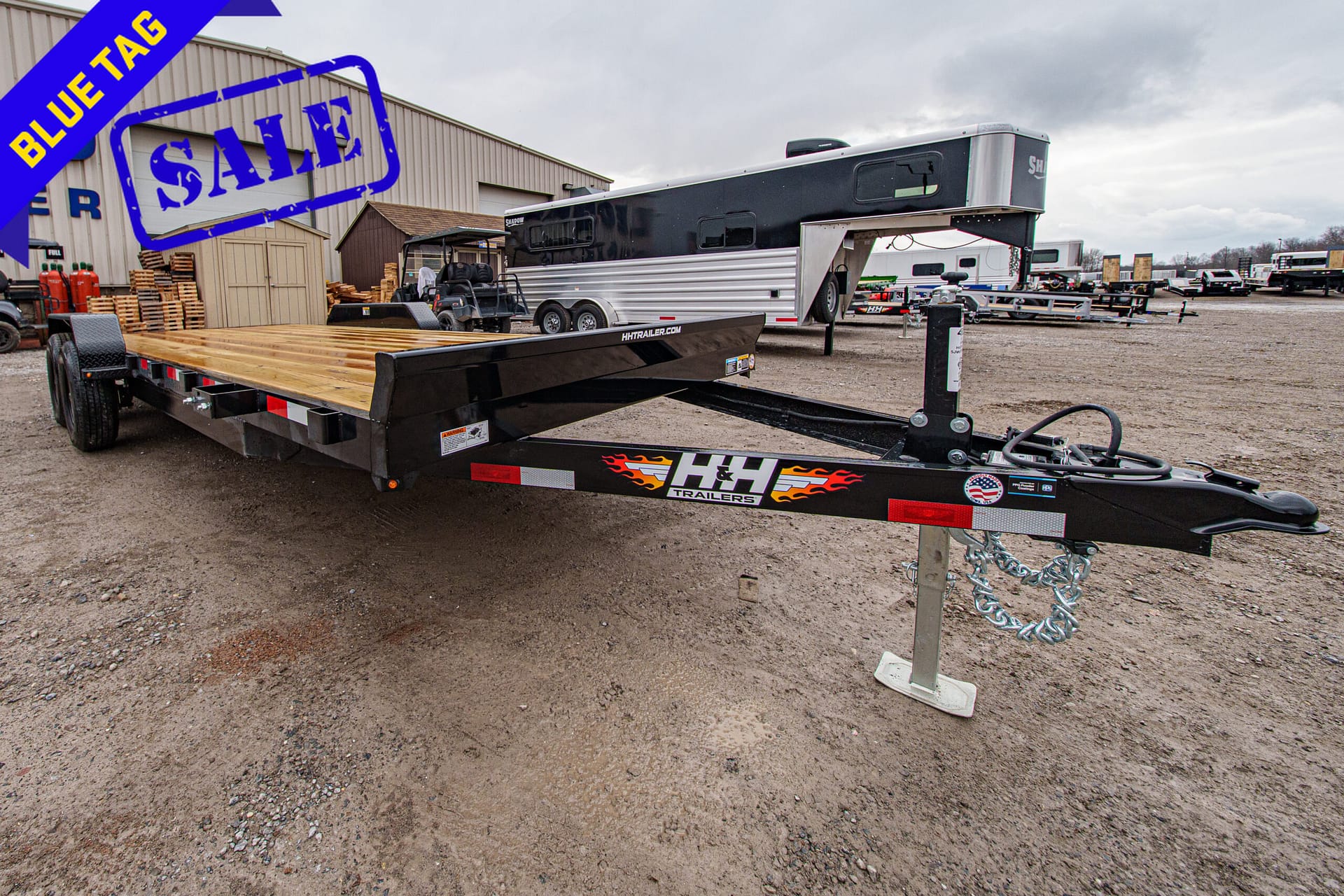 Flatbed trailer in a gravel lot with a large blue 'Blue Tag' sale banner overhead at a trailer yard.