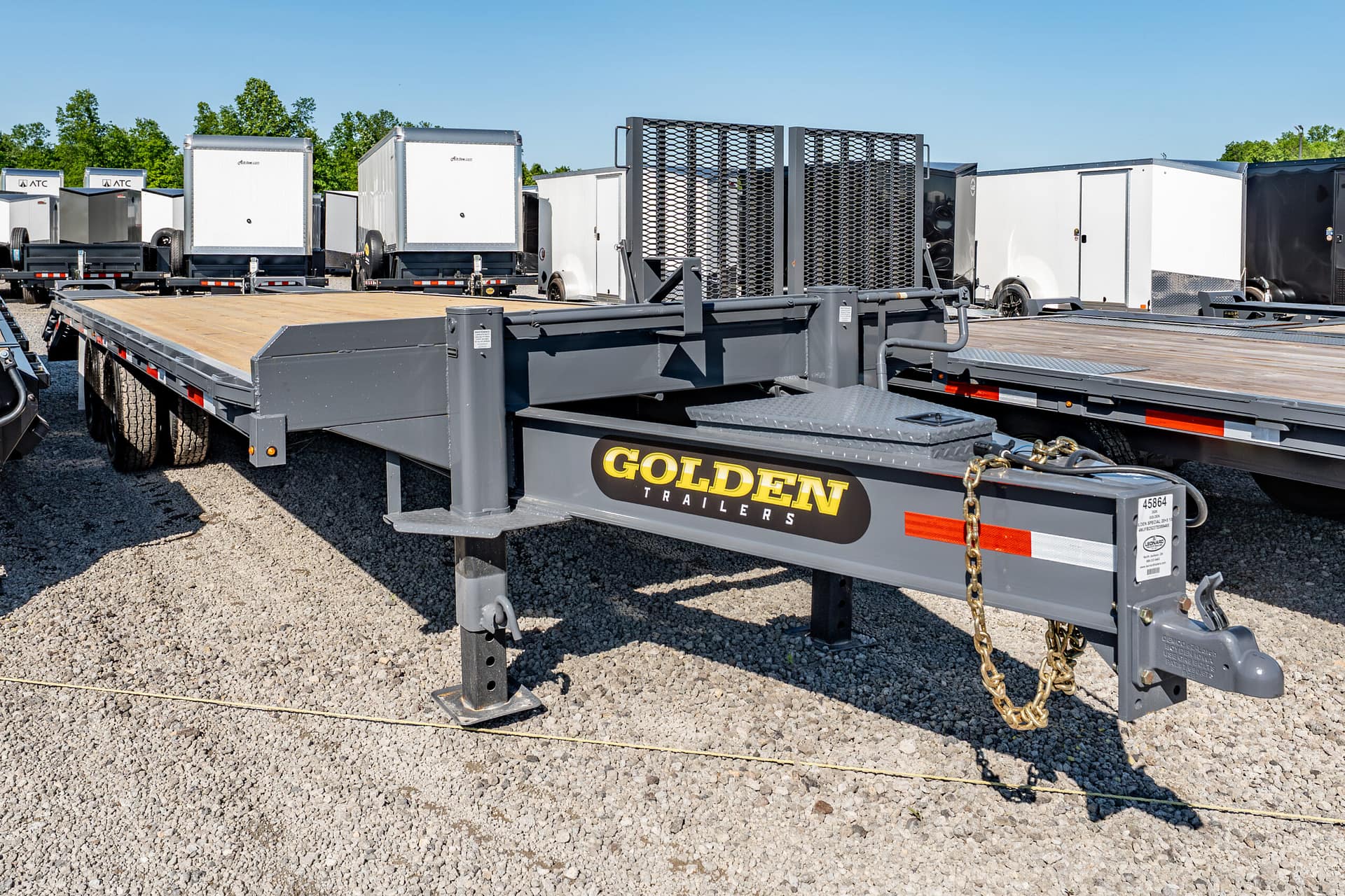 Row of flatbed trailers in a gravel lot, with white utility trailers in the background and a prominent 'Golden Trailers' logo on the foreground trailer.