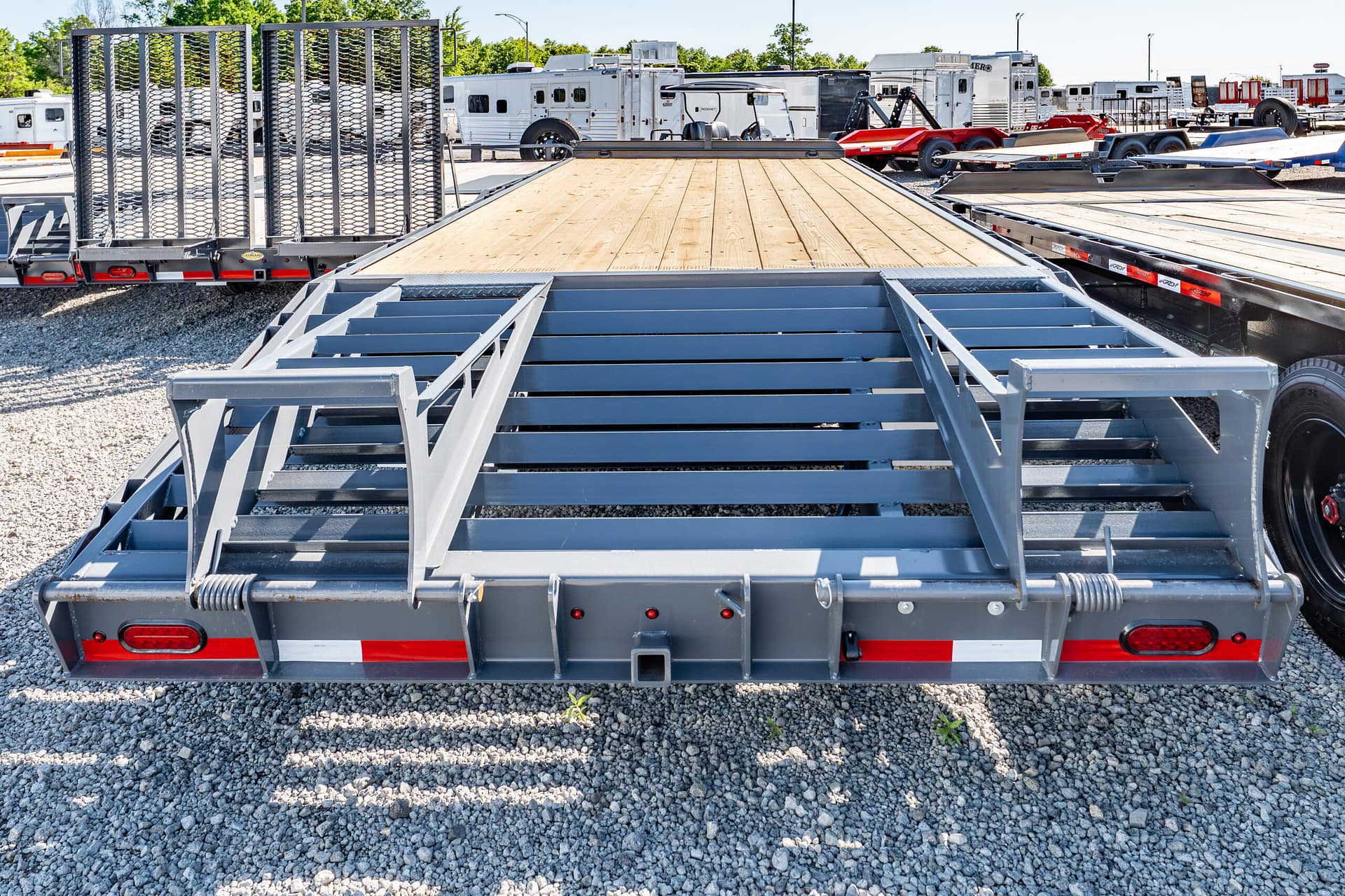 Flatbed trailers with wooden decks parked on a gravel lot, row of equipment in the background, metal framing in foreground