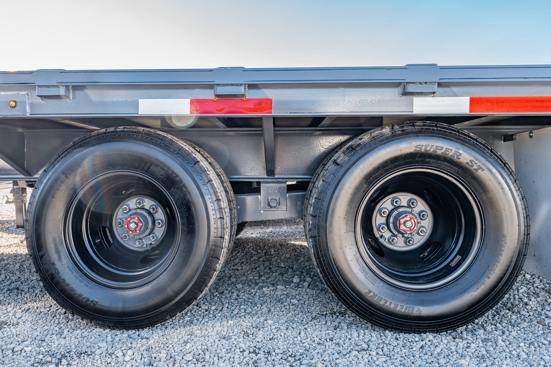 Dual trailer wheels and undercarriage on a gravel surface with reflective striping above the tires.