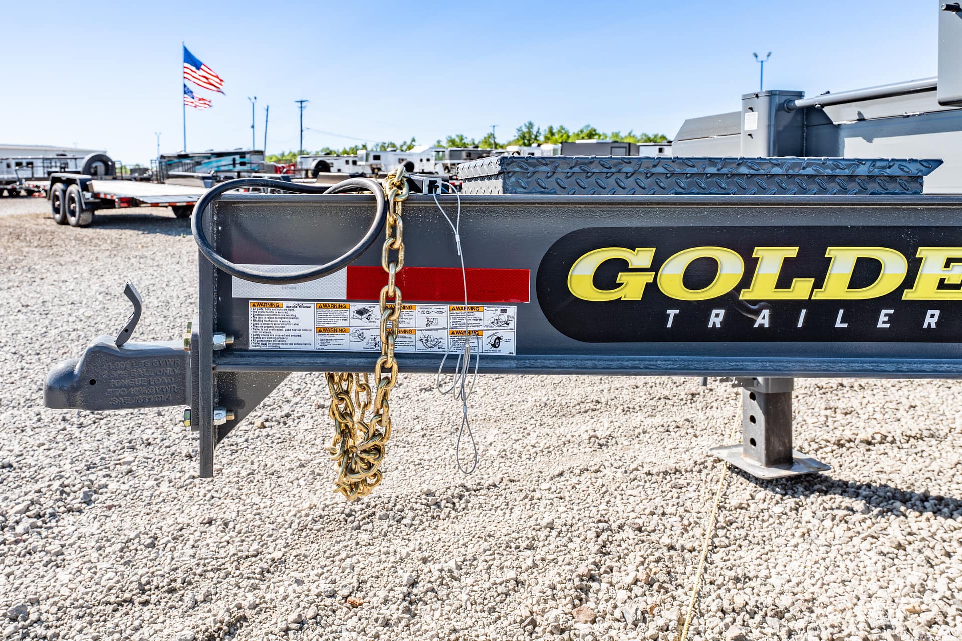 Close-up of a trailer hitch with heavy chains and a diamond-plate toolbox on a gravel lot, with other trailers in the background.