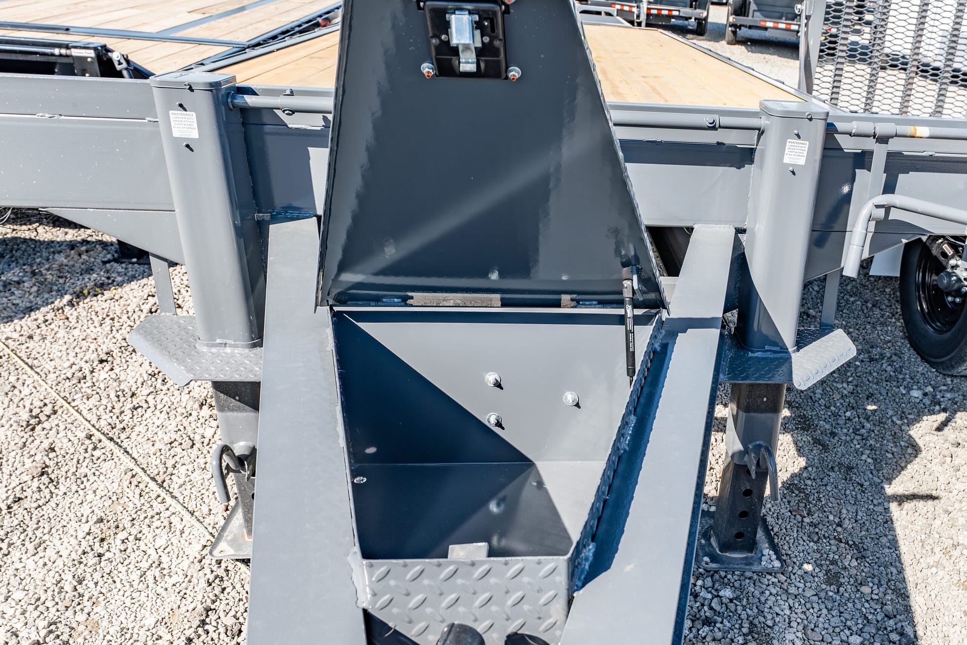 Close-up of a gray metal industrial trailer with a central chute and diamond-plate steps on a gravel surface, likely part of loading equipment.