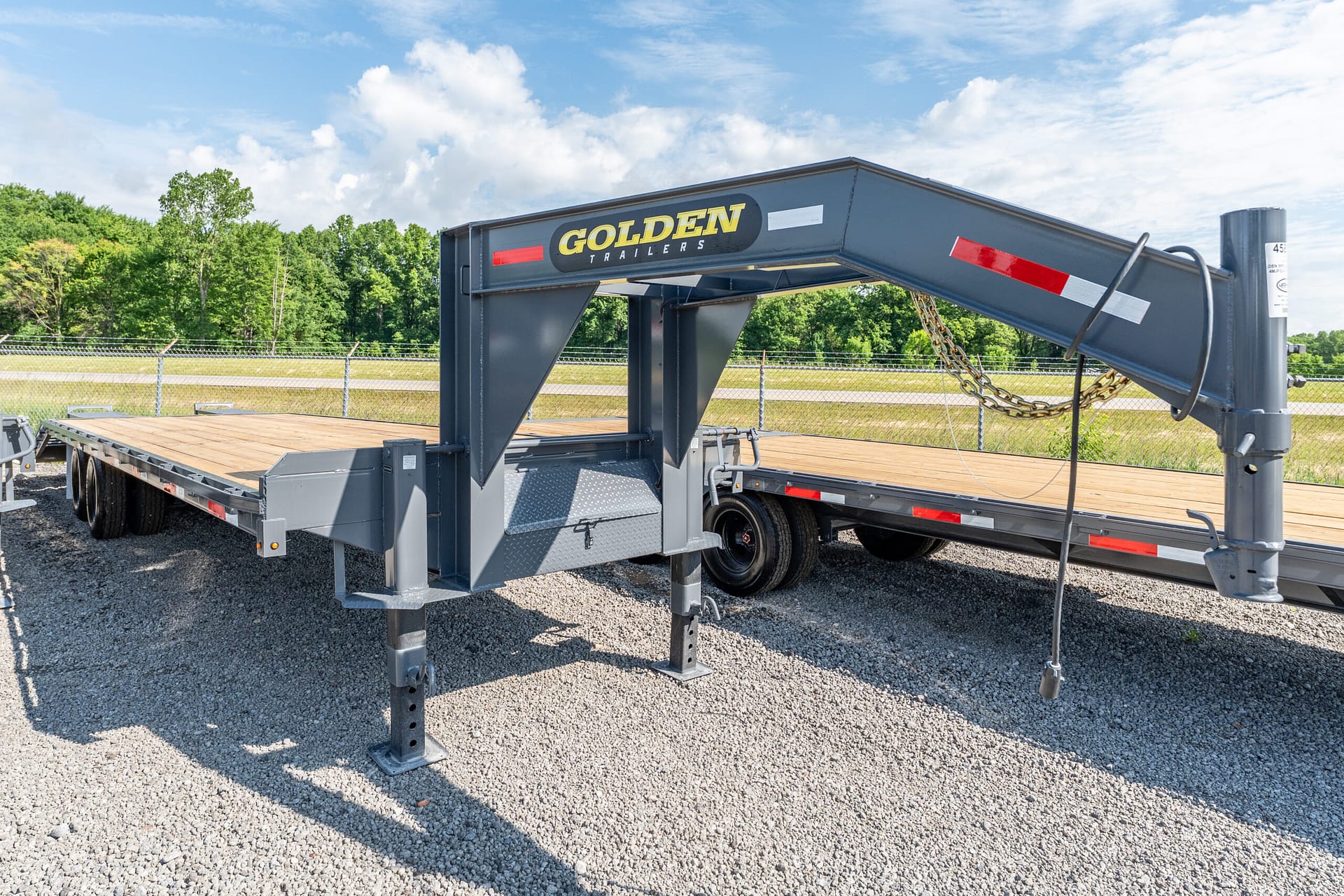 Gooseneck trailer with a gray steel frame and 'GOLDEN TRAILERS' logo in a gravel lot, blue sky and trees in the background.