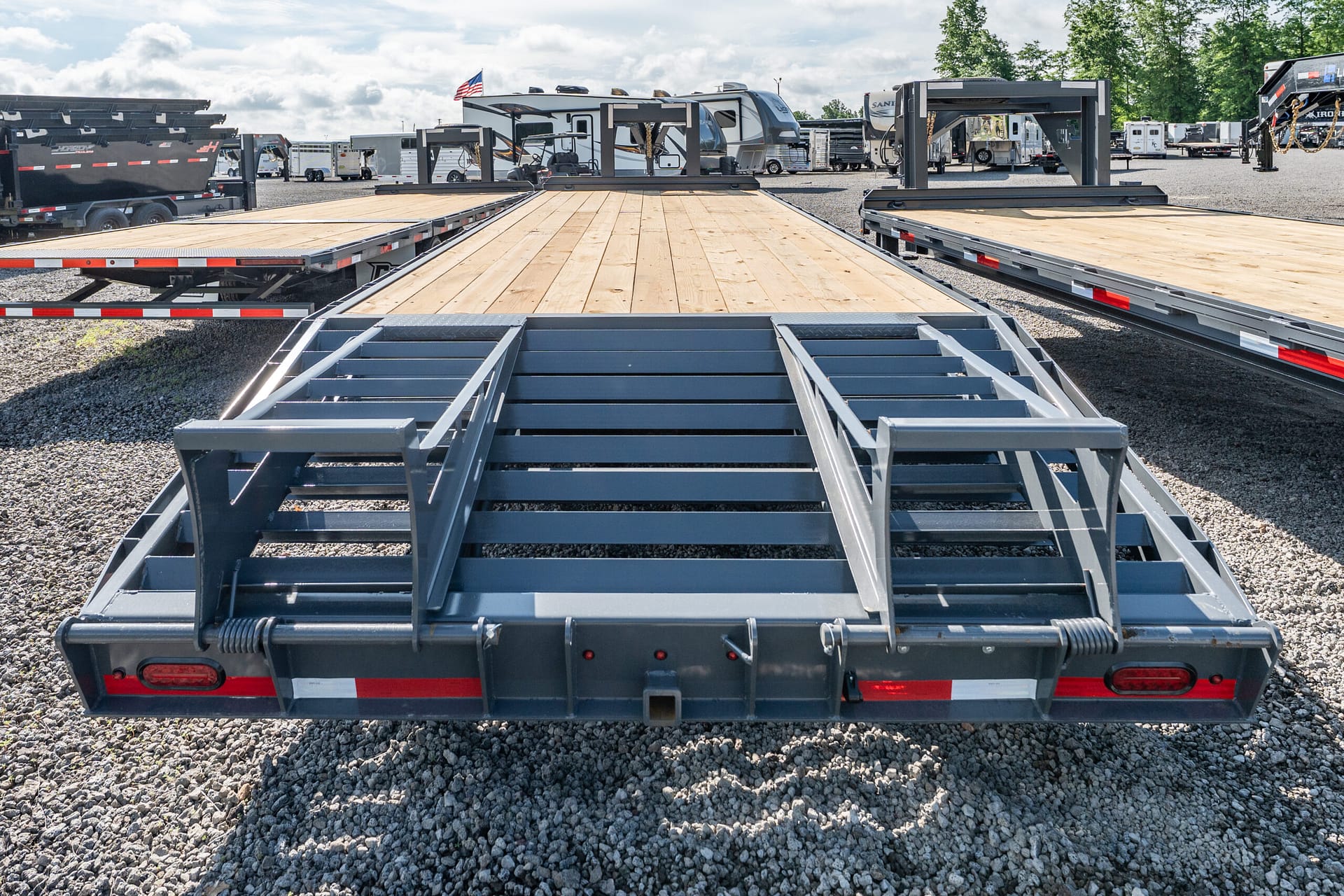 Row of flatbed trailers with wooden decks in a gravel lot, with trucks and RVs visible in the background to indicate a trailer yard.