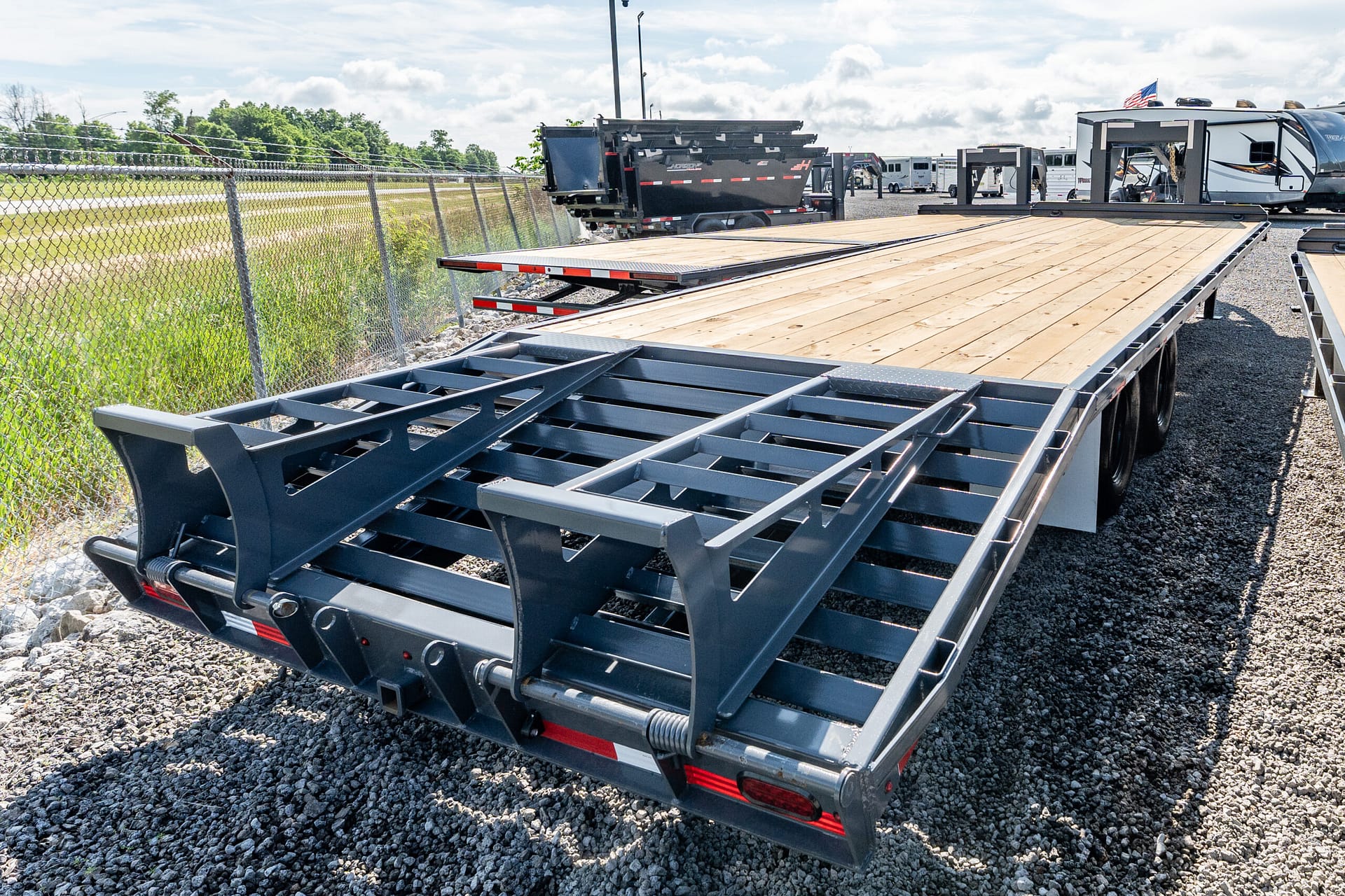 Close view of a metal flatbed trailer with a wooden deck on a gravel lot, next to a chain-link fence.