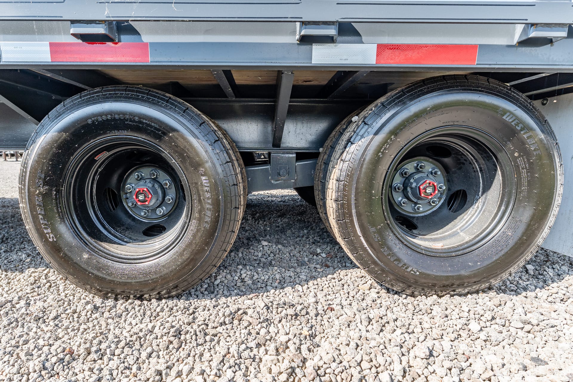 Dual trailer wheels and undercarriage with red hub covers, resting on a gravel surface.