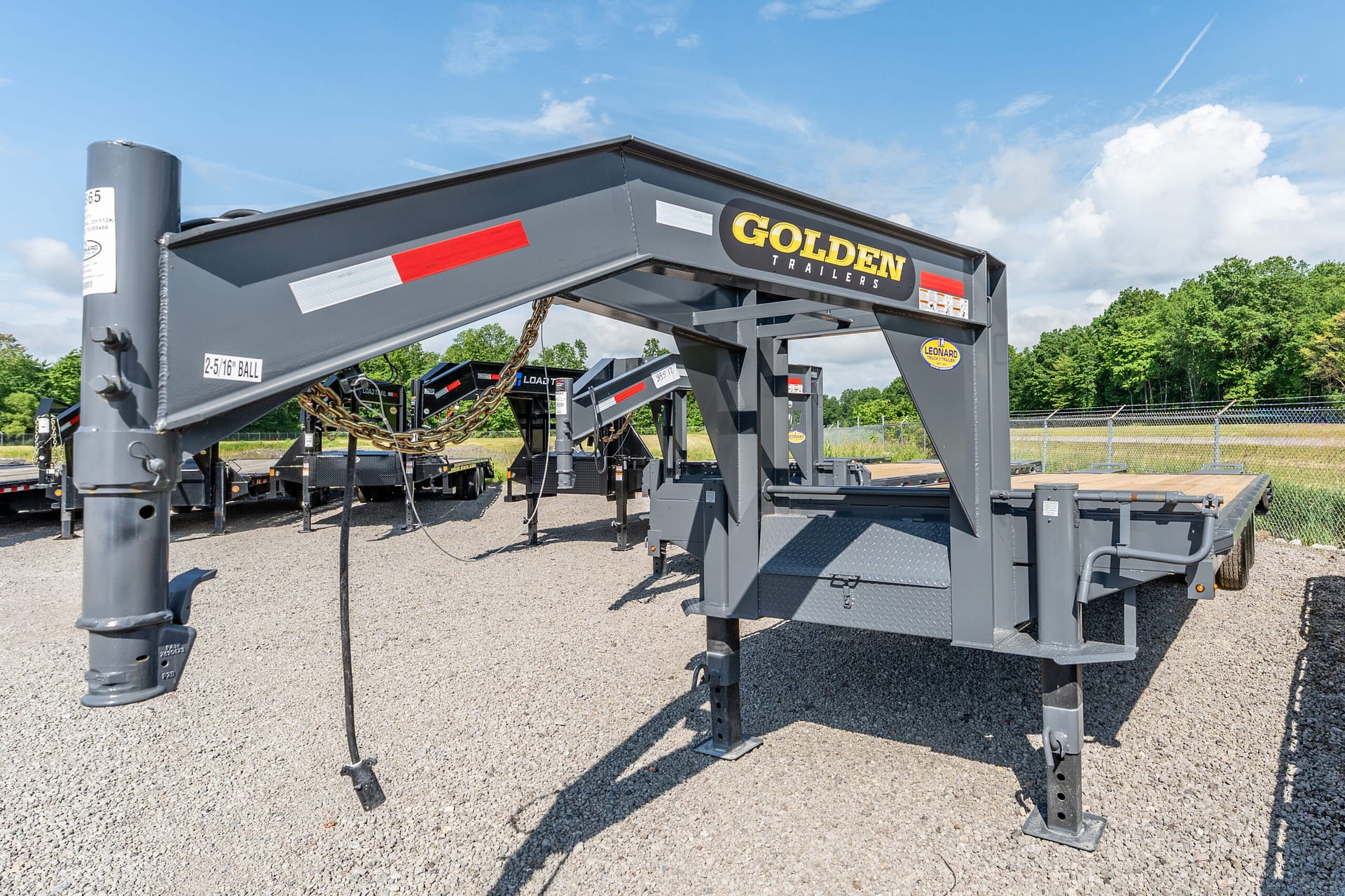 Grey industrial trailer frame with a chain and hitch, featuring a prominent 'GOLDEN TRAILERS' logo, parked on a gravel lot with a fence and trees in the background.