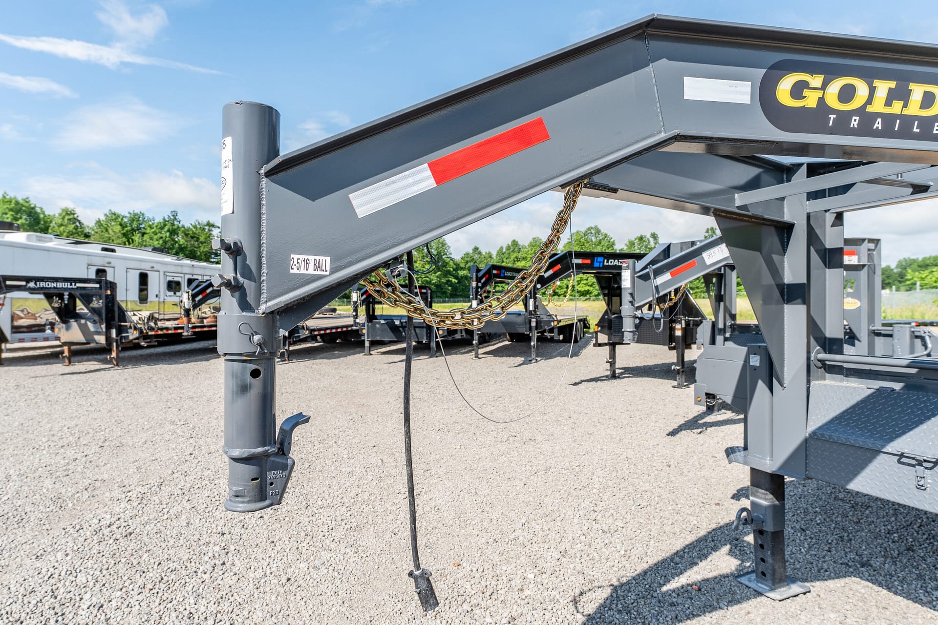 Close-up of a gray industrial trailer tongue with red/white reflective strips, chains, and support beams in a gravel lot.