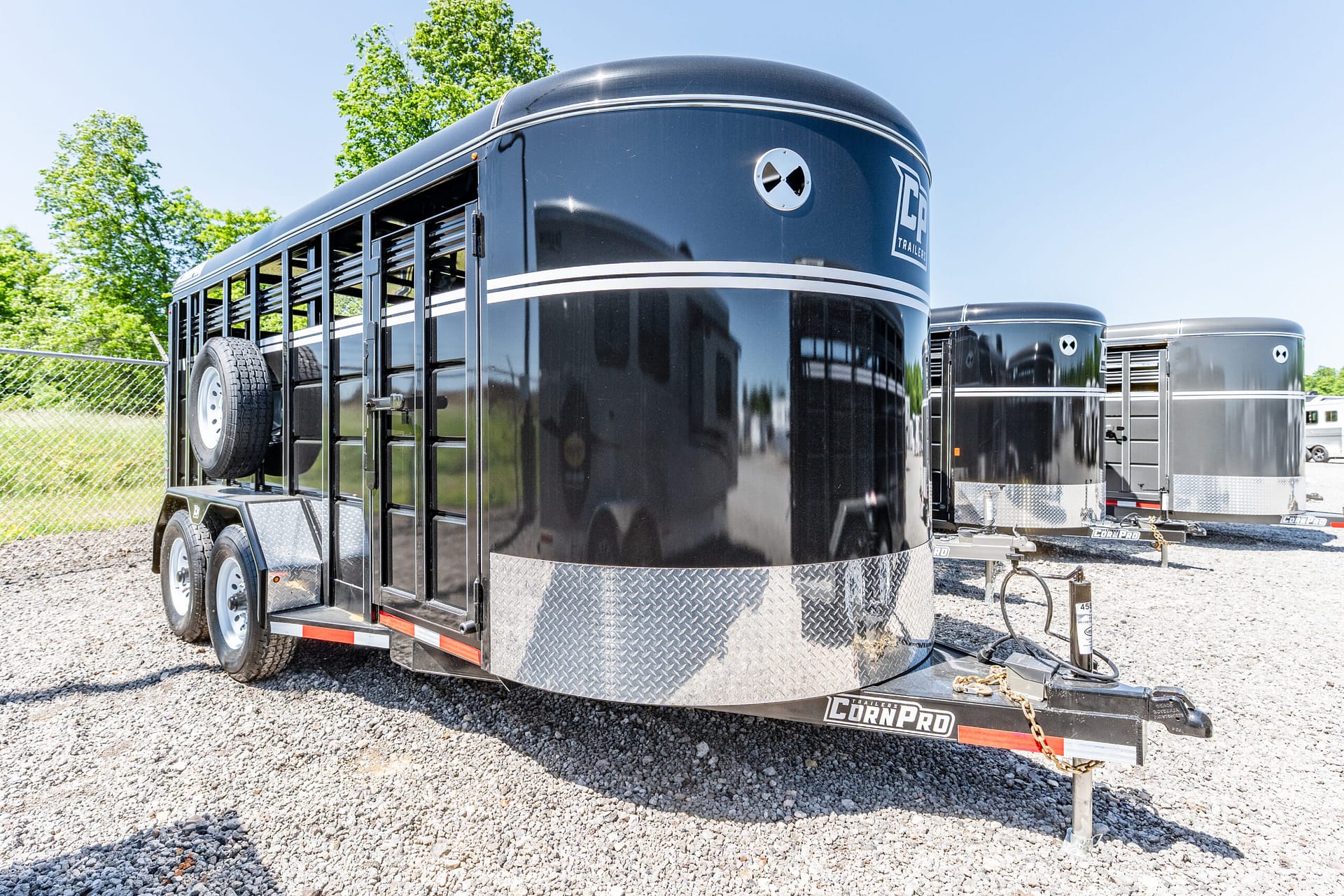Black enclosed trailer with a white stripe and chrome diamond-plate trim, spare tire mounted on the side, on a gravel lot with other trailers in the background.