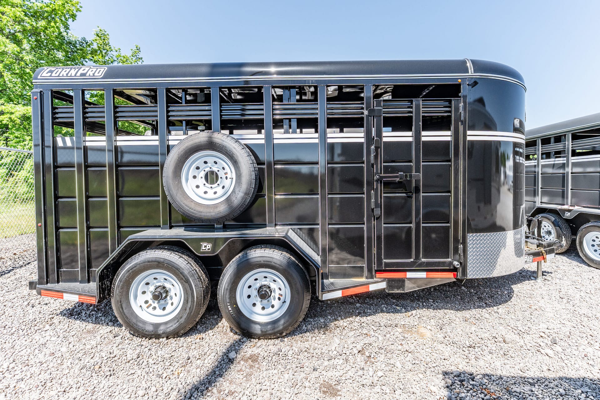Black CornPro livestock trailer with a spare tire mounted on the side, parked on a gravel lot under a clear blue sky, trees in the background.