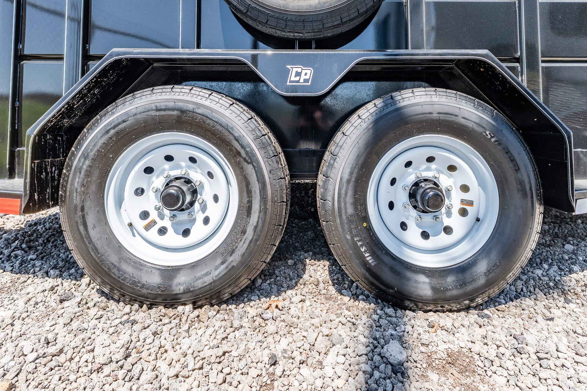 Dual-axle trailer wheels with a spare tire mounted above on a black metal frame, parked on gravel.