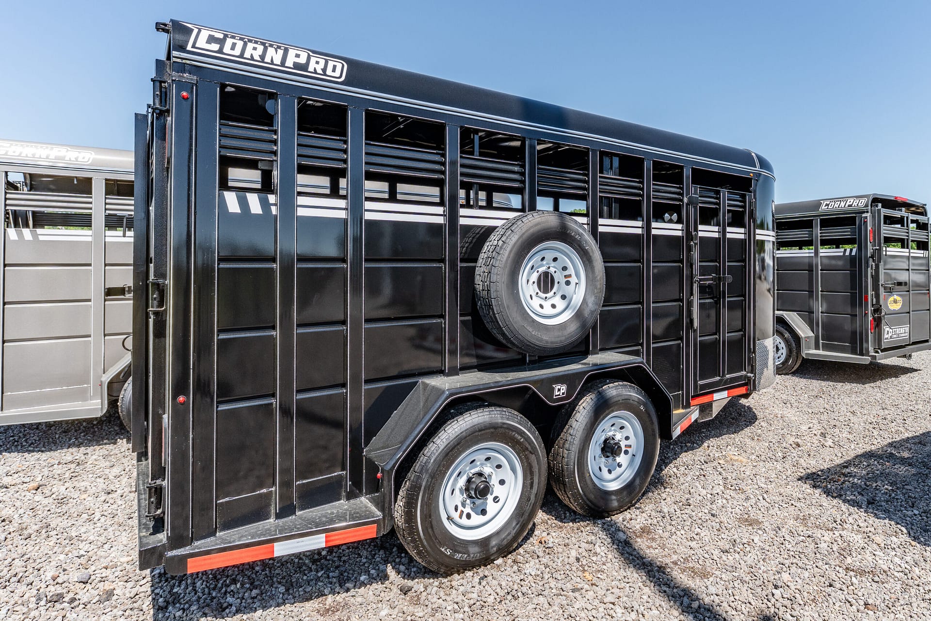 Black livestock trailer with a side-mounted spare tire, parked on gravel under a clear blue sky.