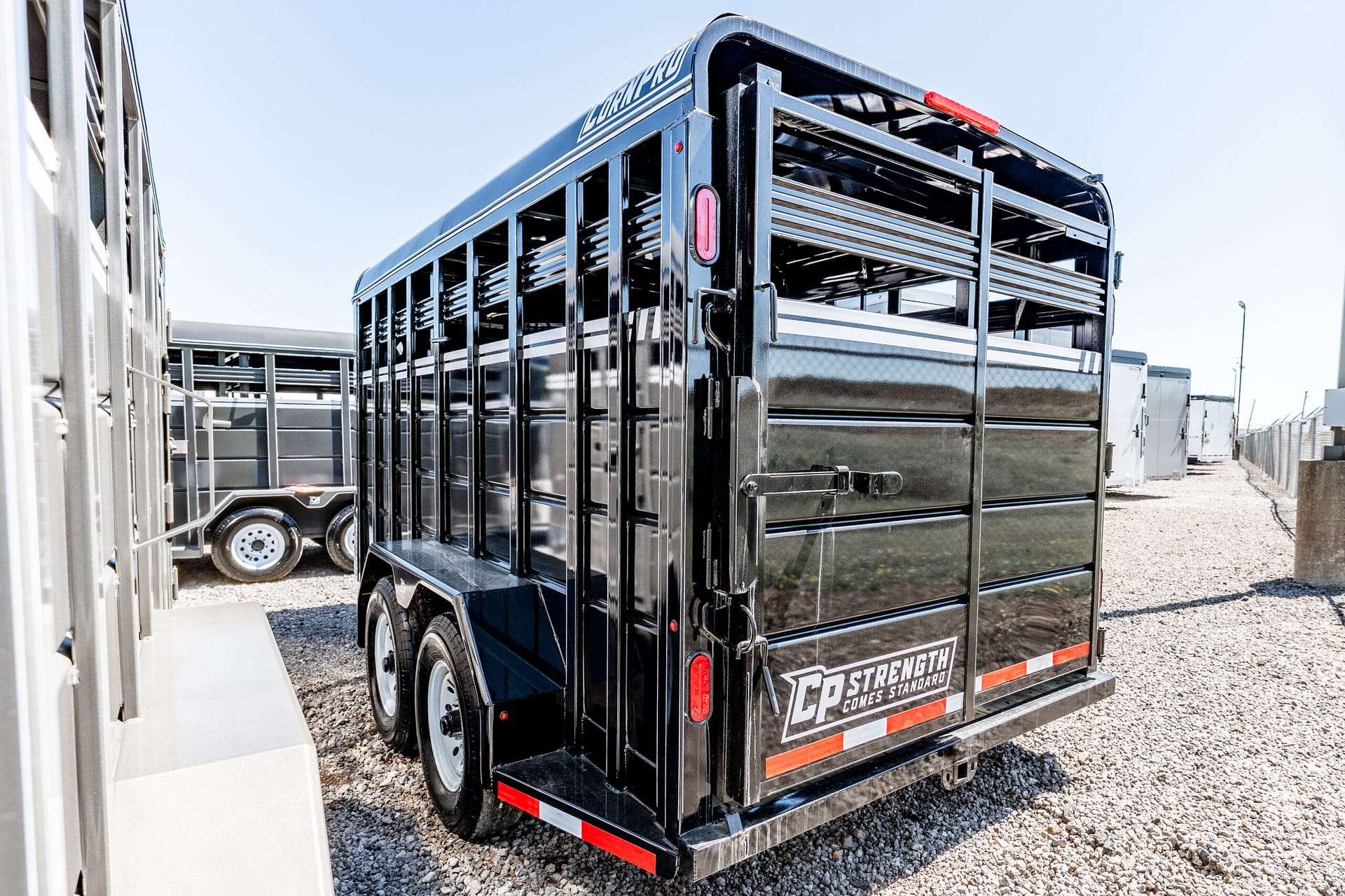 Rear view of a glossy black livestock trailer with red taillights and chrome rails in a gravel lot, with other trailers in the background.