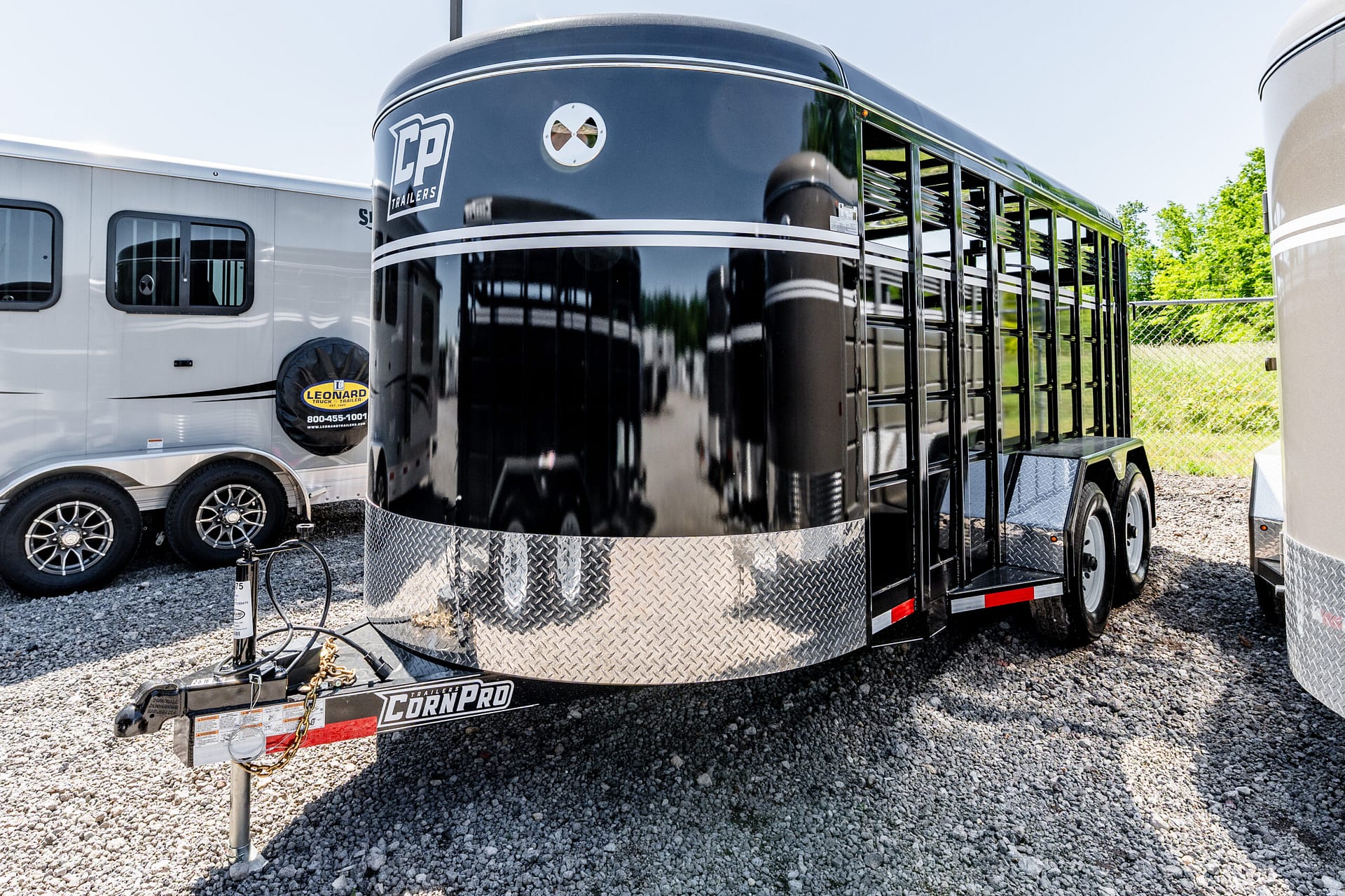 Close-up of a glossy black livestock trailer with a diamond-plate lower front, hitch and safety chain on a gravel lot, beside a white trailer.