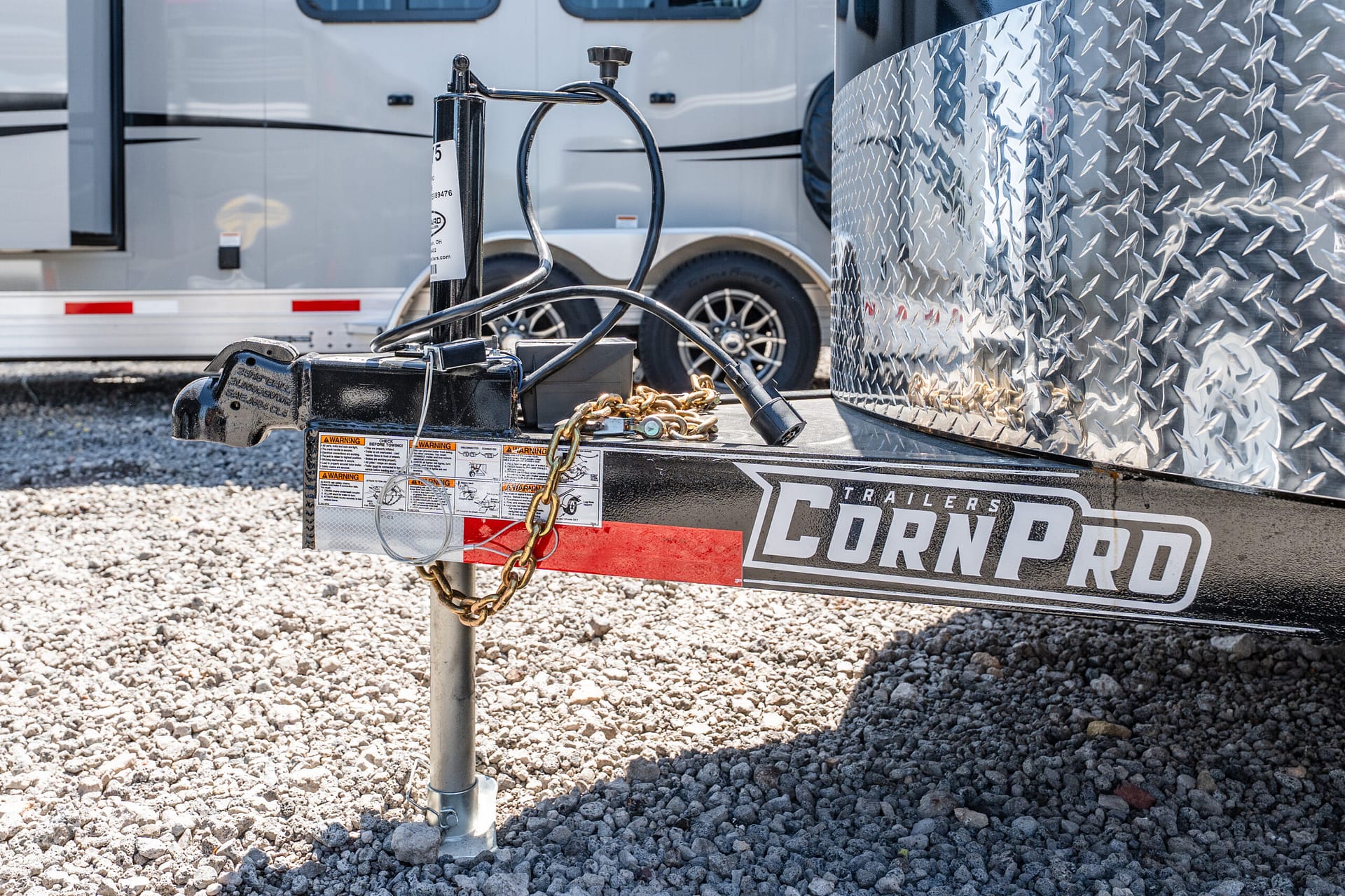 Close-up of a trailer hitch with safety chains and wiring on a gravel lot, with a diamond-plate trailer wall in the background.