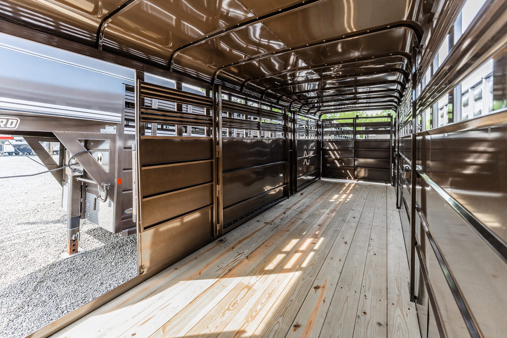 Interior of a brown aluminum livestock trailer with partitioned stalls and a wooden floor, sunlight filtering in from side windows.