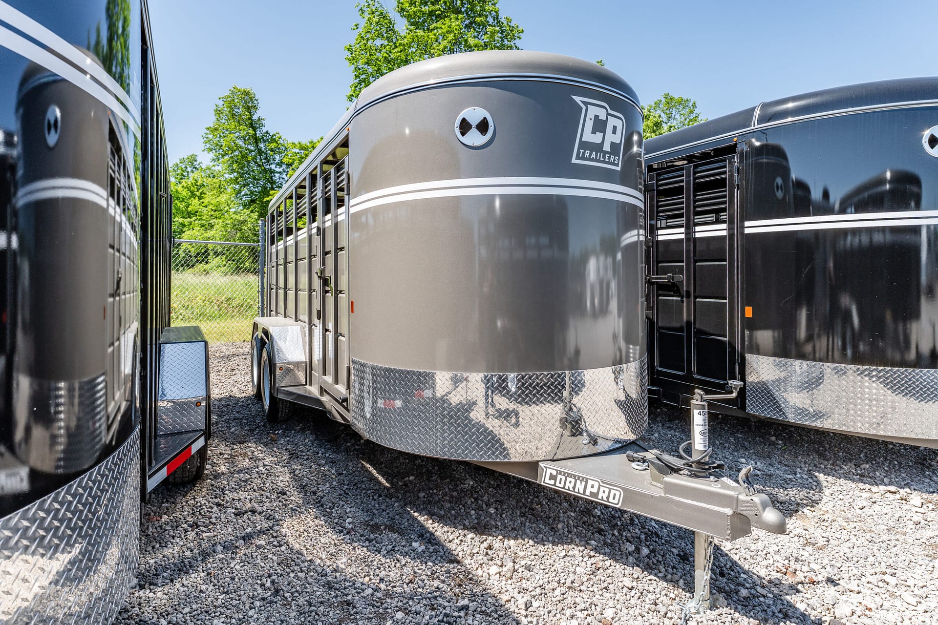 Grey cattle trailer with white stripe parked between other trailers on a gravel lot, green trees in the background.
