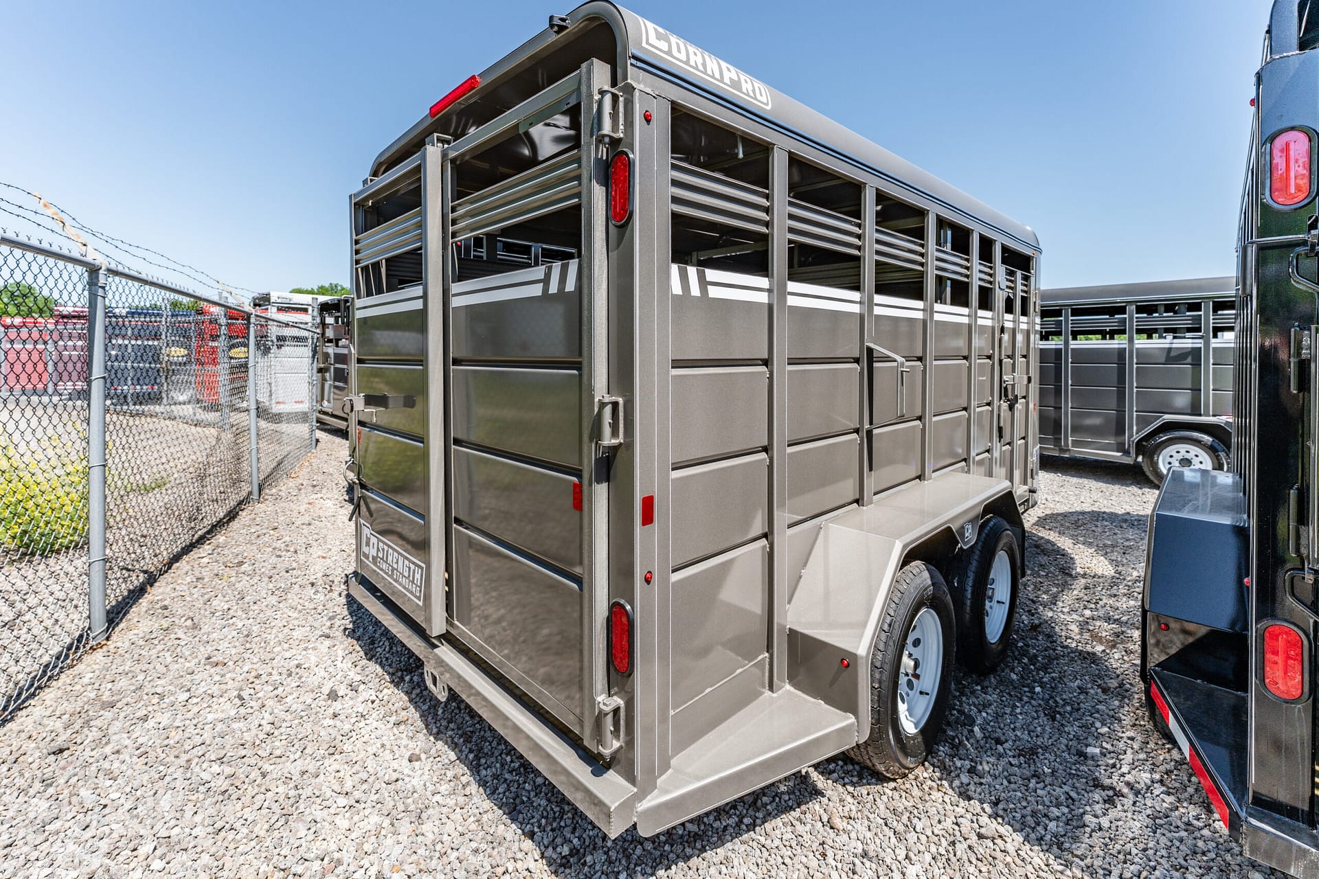 Gray metal livestock trailer with slatted sides, parked on a gravel lot beside a chain-link fence under a clear blue sky.