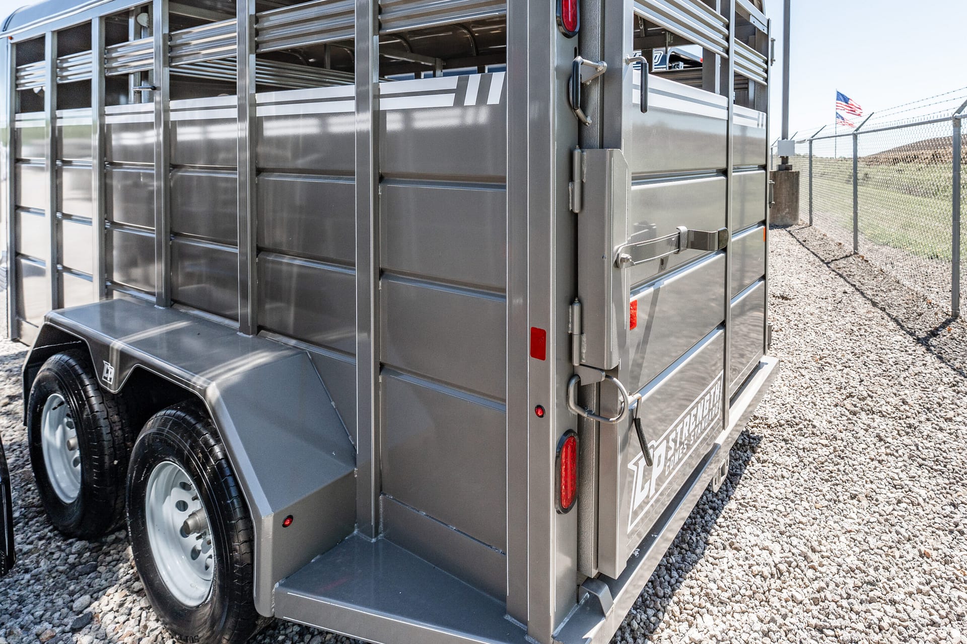 Close-up of a silver livestock trailer with ribbed metal sides and dual rear doors on a gravel lot; fence and an American flag visible in the background.