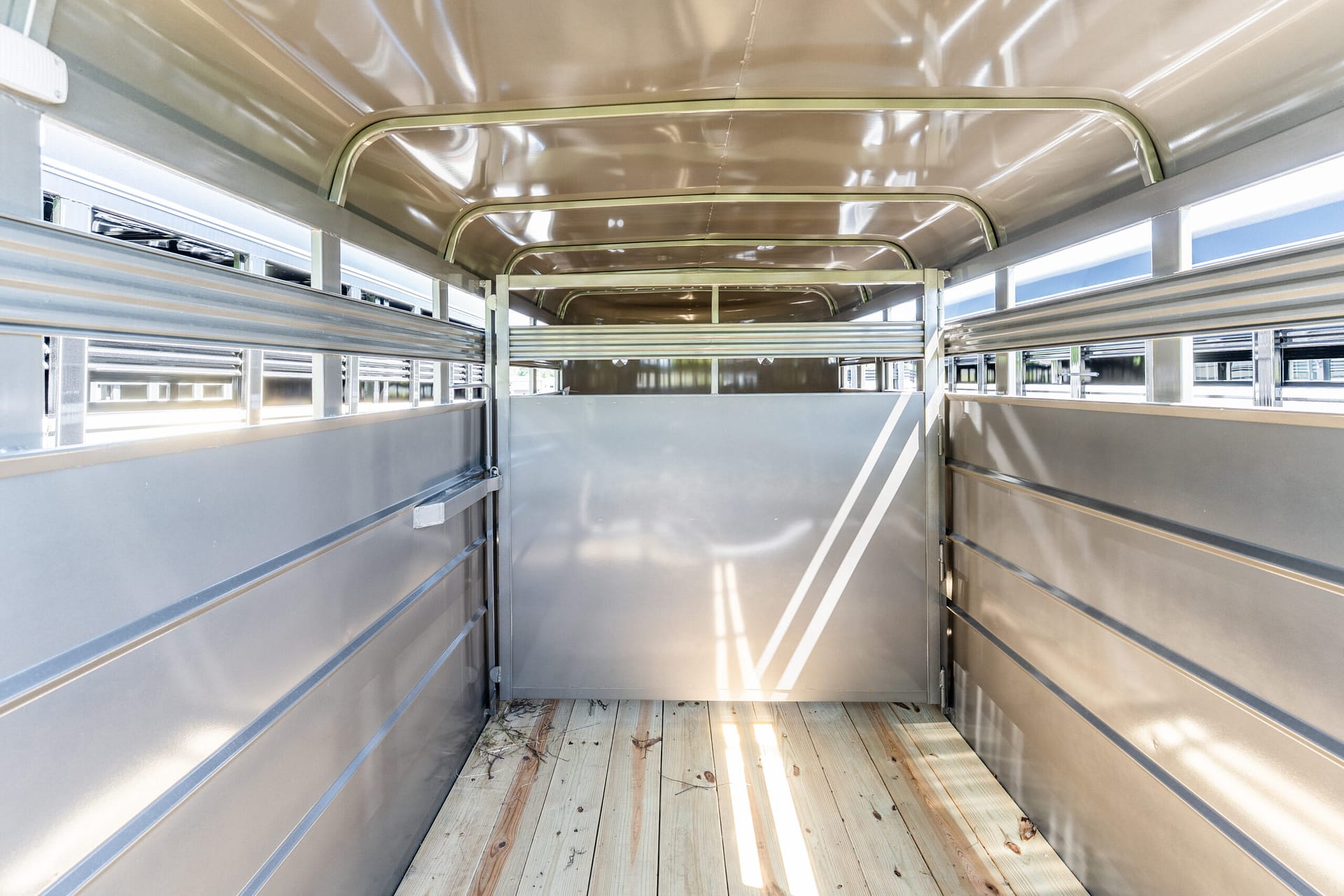 Inside an empty stainless steel trailer with side rails and a wooden floor, viewed from the back aisle to the front end.