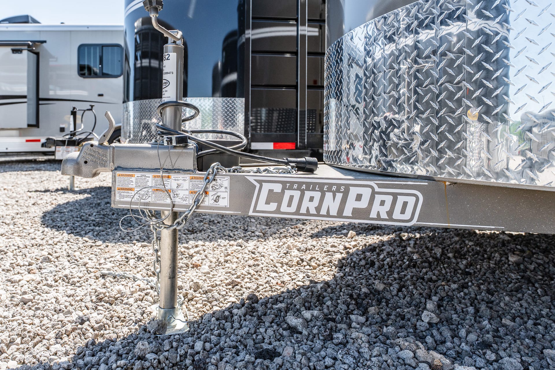 Close-up of a trailer hitch and stabilizing jack on a gravel lot, with a diamond-plate trailer body nearby.
