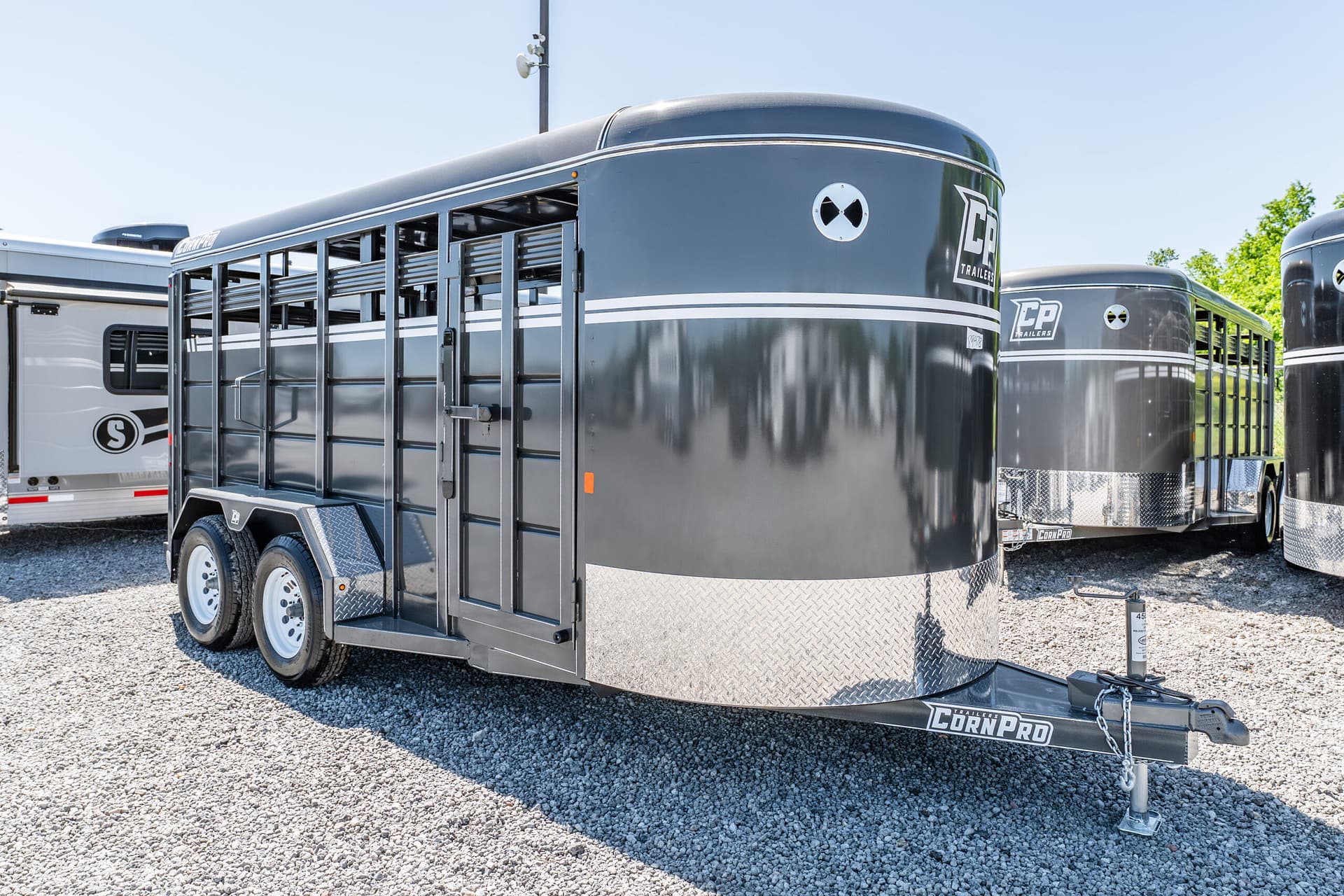 Gray livestock trailer with vented side doors and a CornPro logo, parked on gravel with other trailers nearby