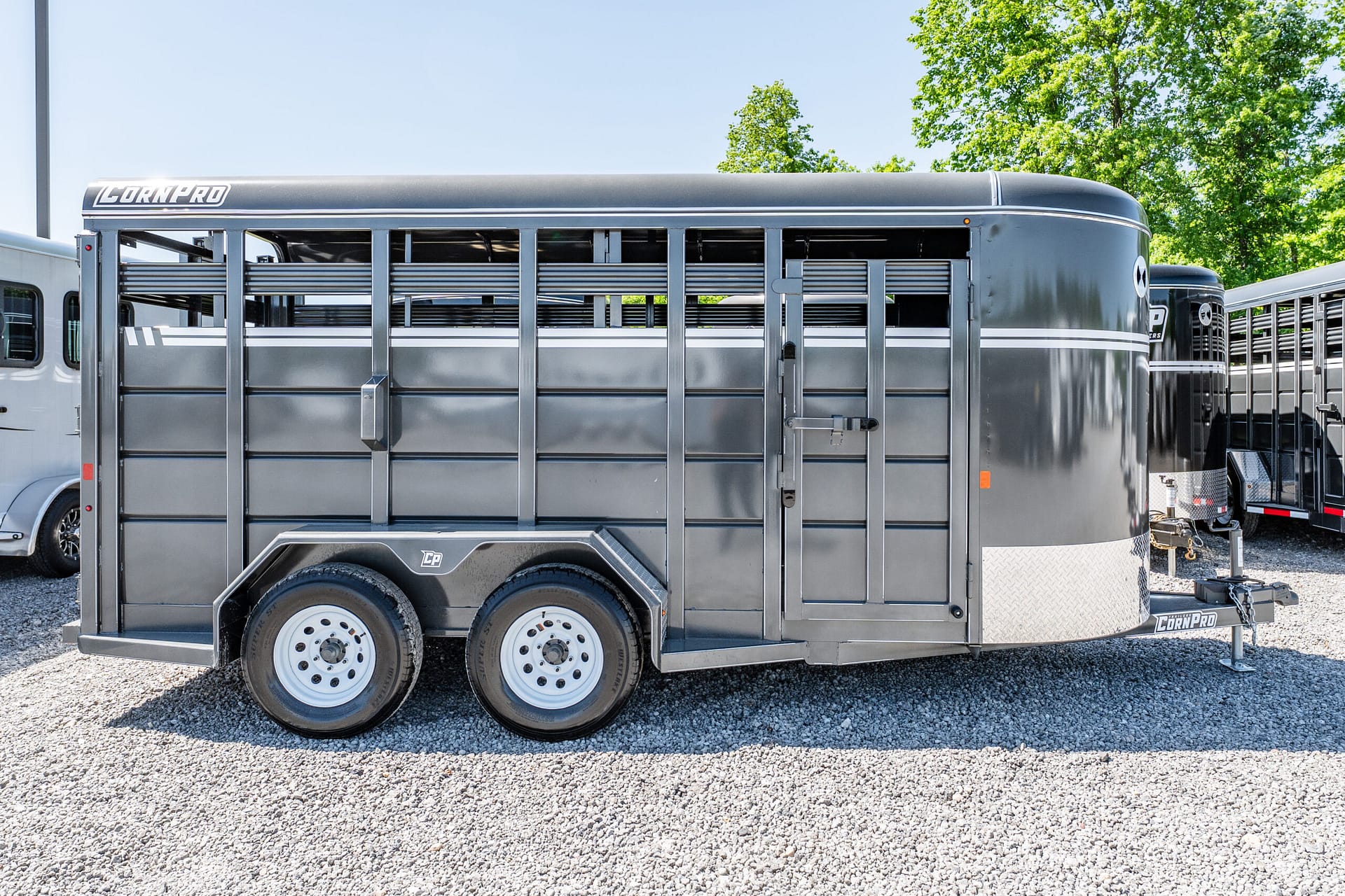 Gray livestock trailer with vented side slats, parked on a gravel lot with green trees in the background