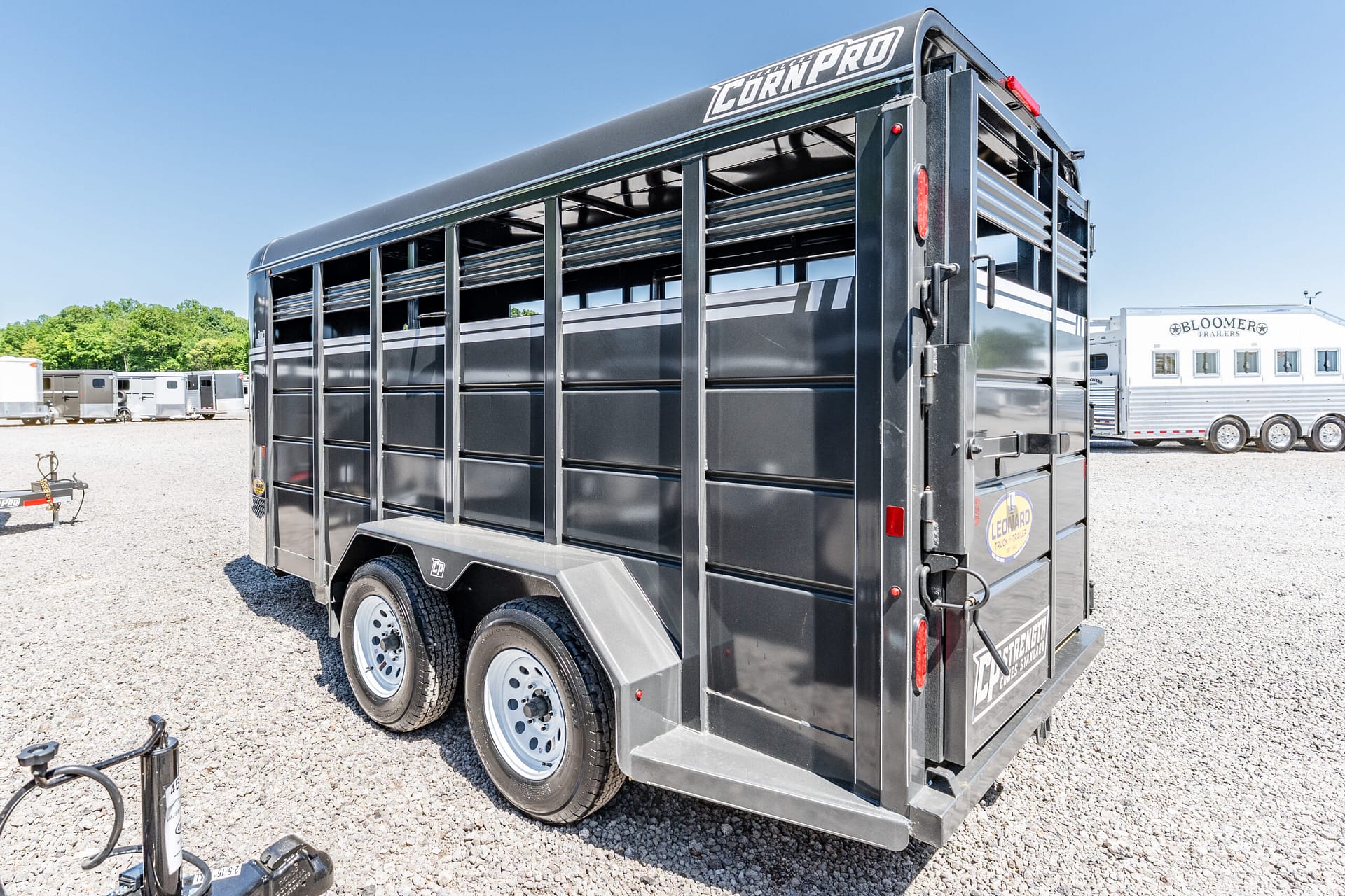 Black livestock trailer with ventilated side panels and dual axles, parked on a gravel lot with other trailers and a clear blue sky in the background.