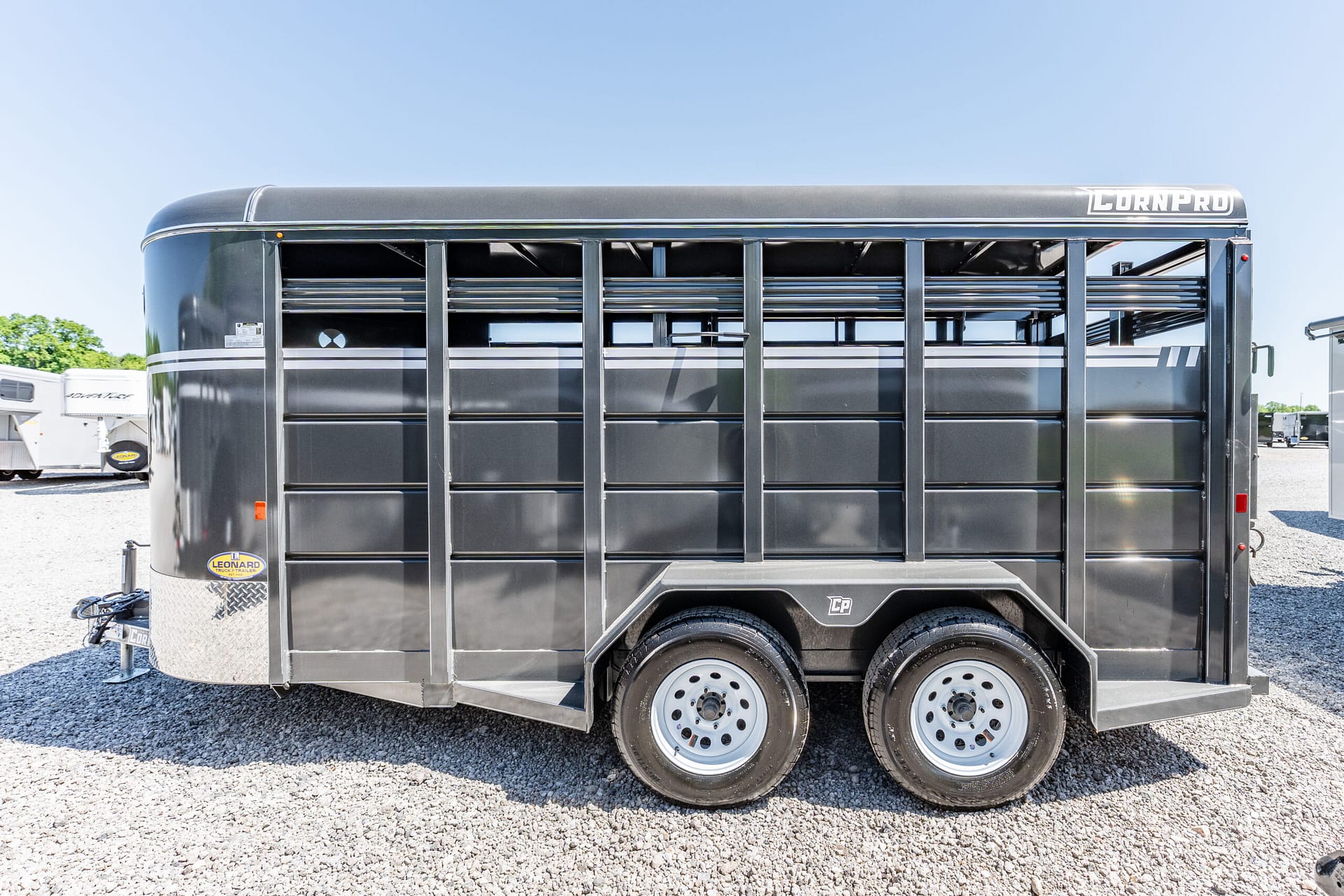 Black livestock trailer with ventilation slats and two axles parked on gravel, front-right side visible.