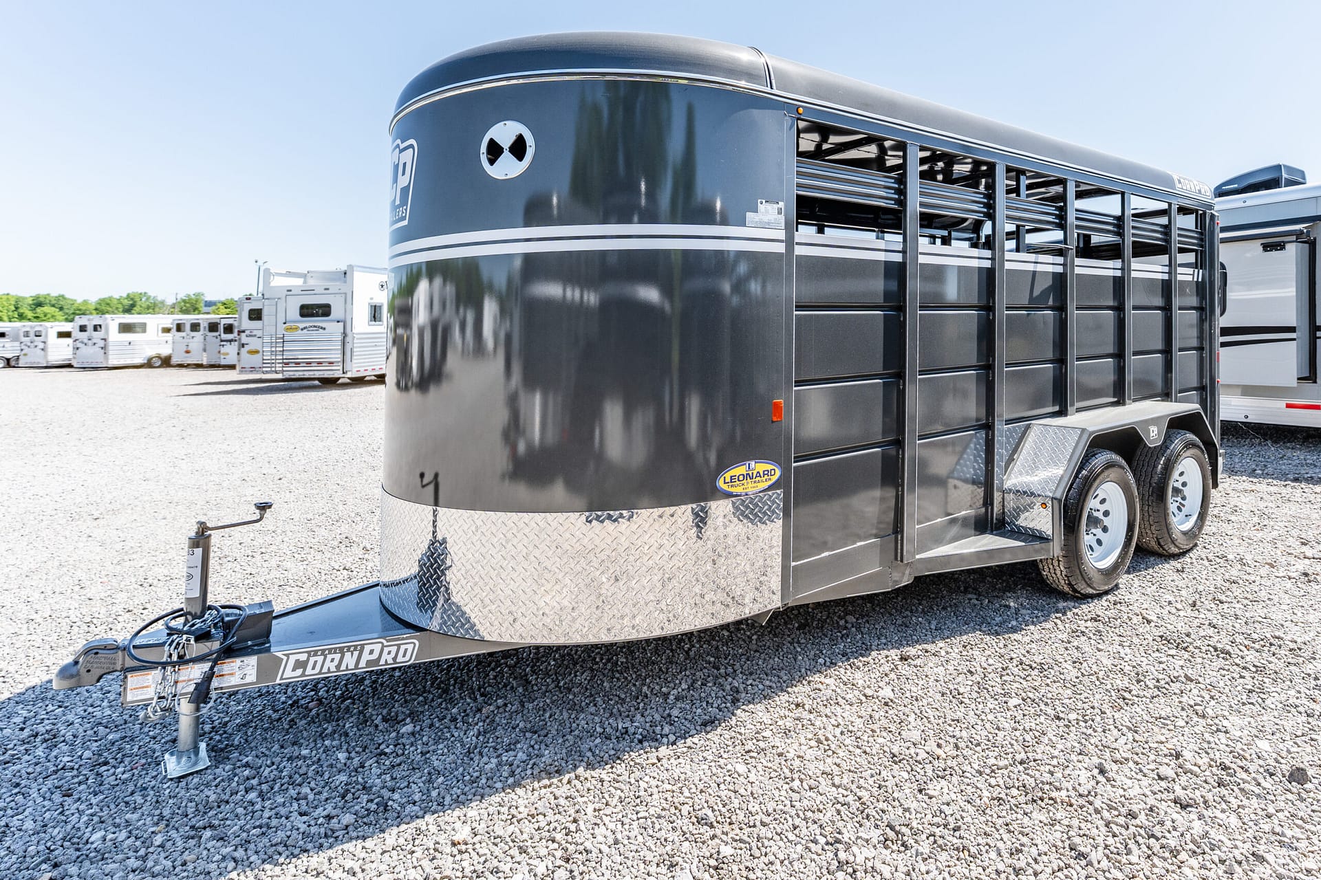 Close-up of a dark livestock trailer with white stripes, parked on a gravel lot with other trailers in the background.
