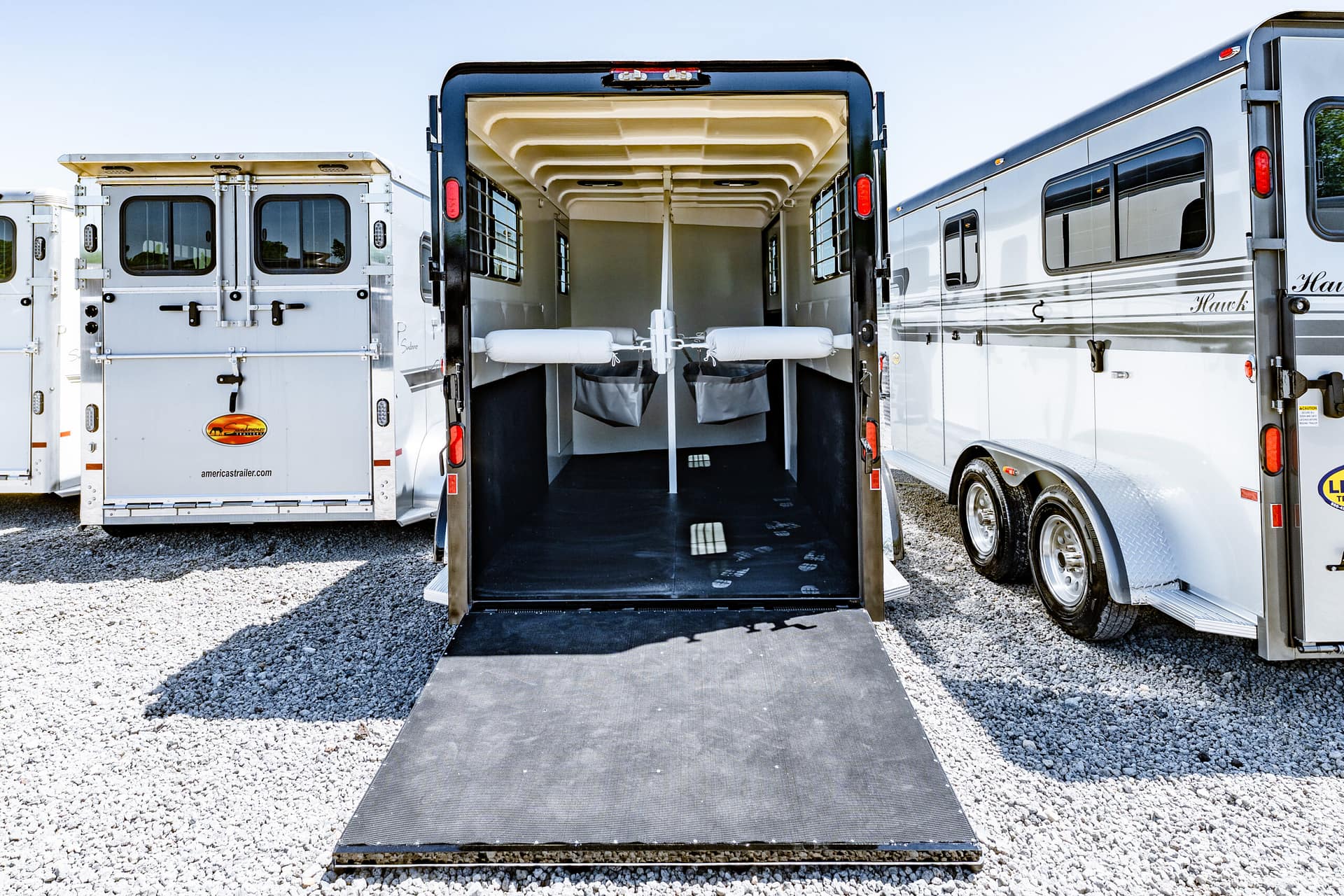 Open rear of a black horse trailer with its ramp down, revealing interior stalls and tie rails on a gravel lot between other trailers.