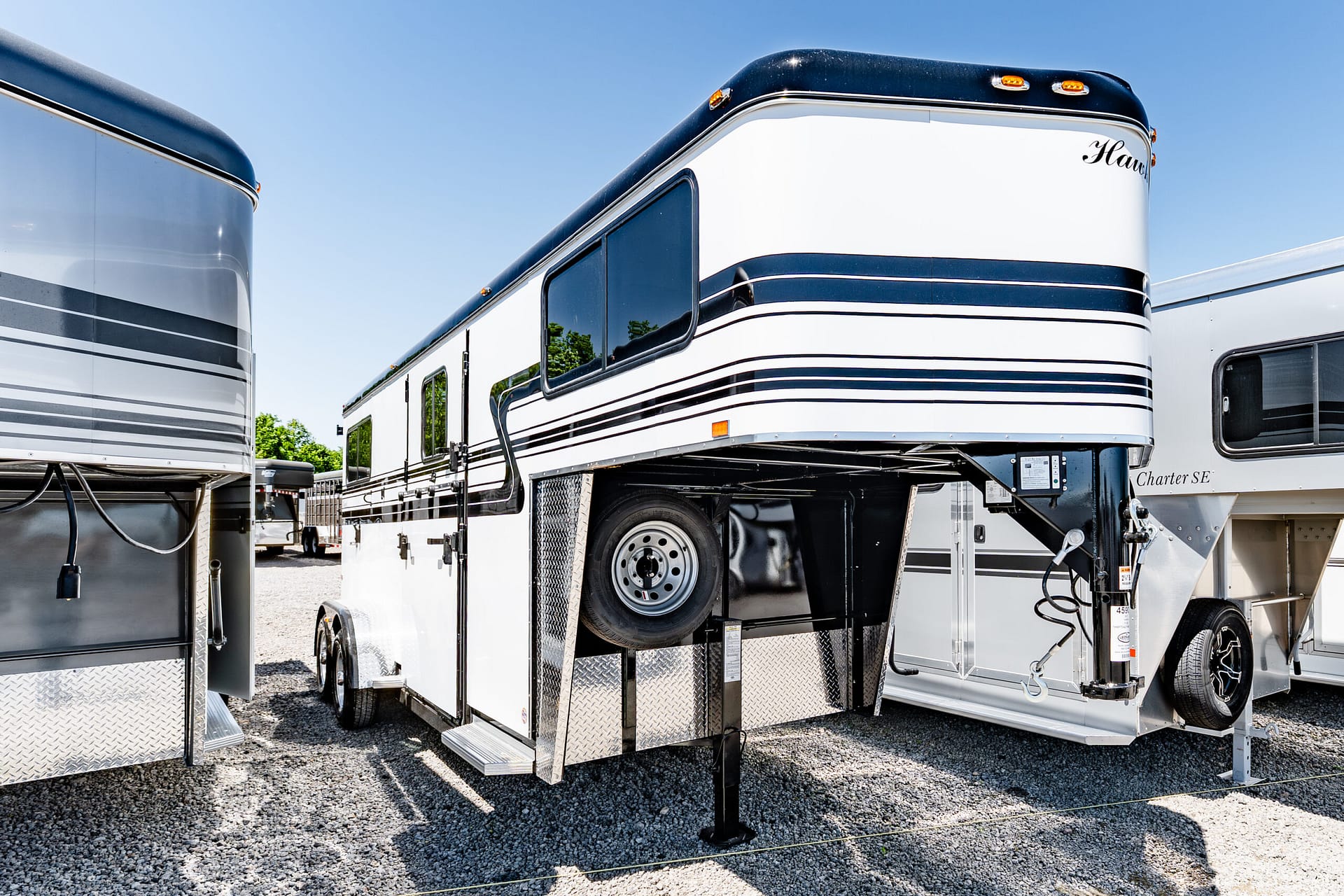 White and blue fifth-wheel trailer raised on jacks in a gravel lot, with another trailer visible in the background.