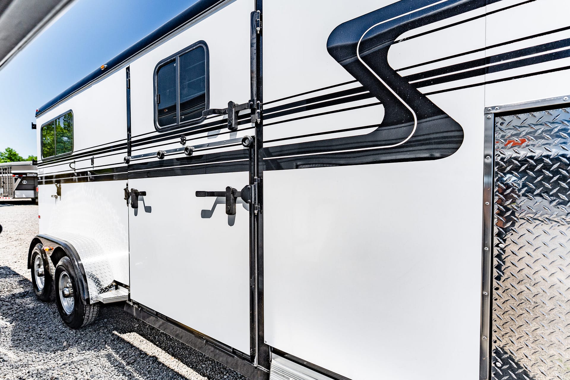 White trailer with black decorative stripes, steel latches, and diamond-plate panel at the rear on a gravel lot.