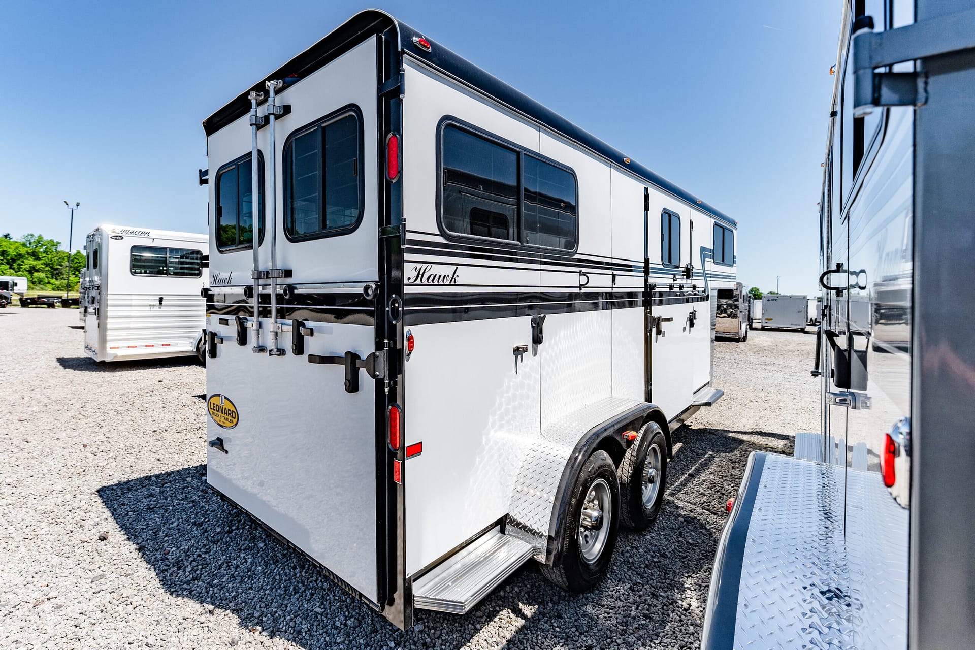 Rear corner of a white Hawk travel trailer with black stripes, parked on gravel with other trailers in the background under a clear blue sky.