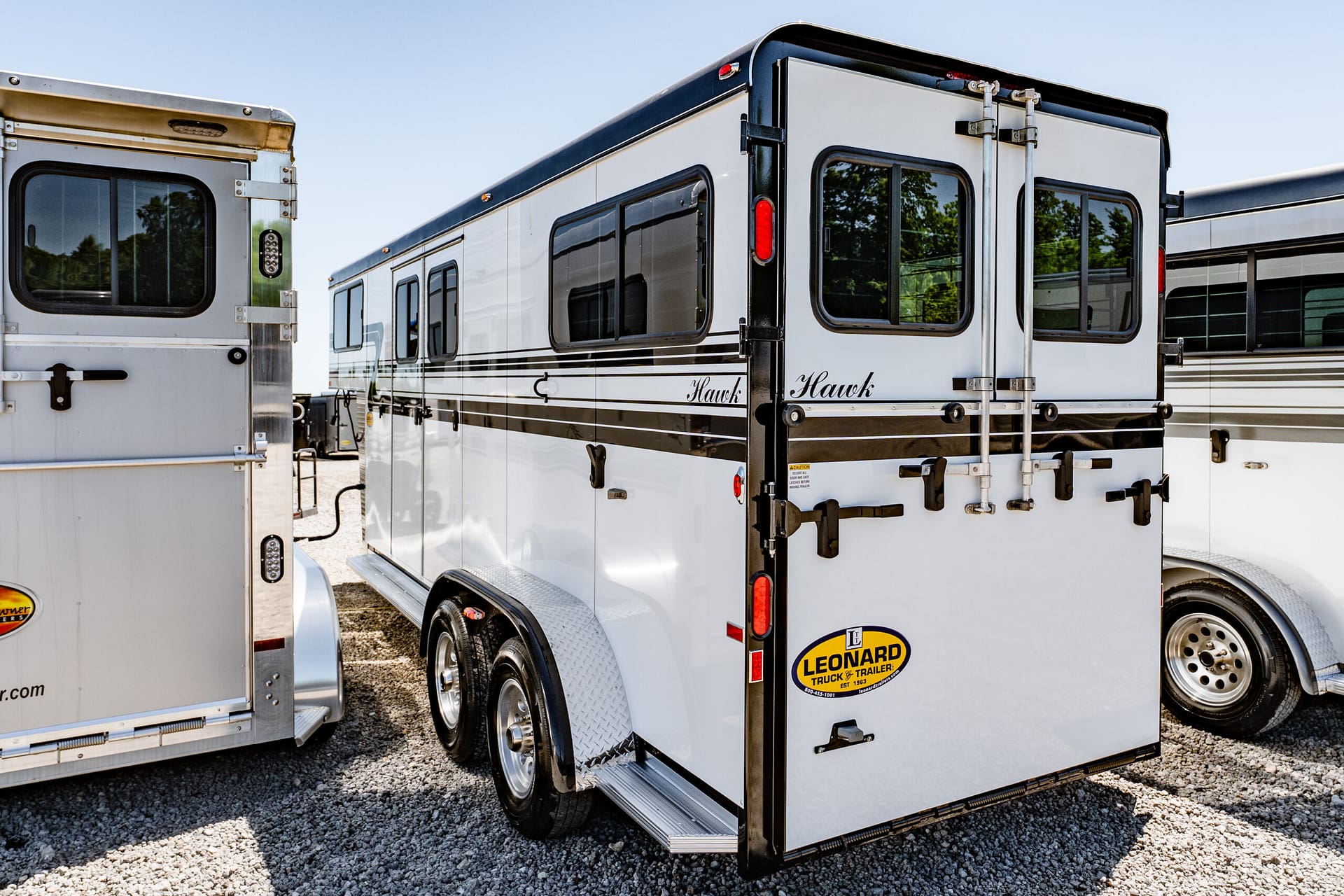 White Hawk horse trailer with black stripes and red taillights parked on gravel yard, side and rear shown.