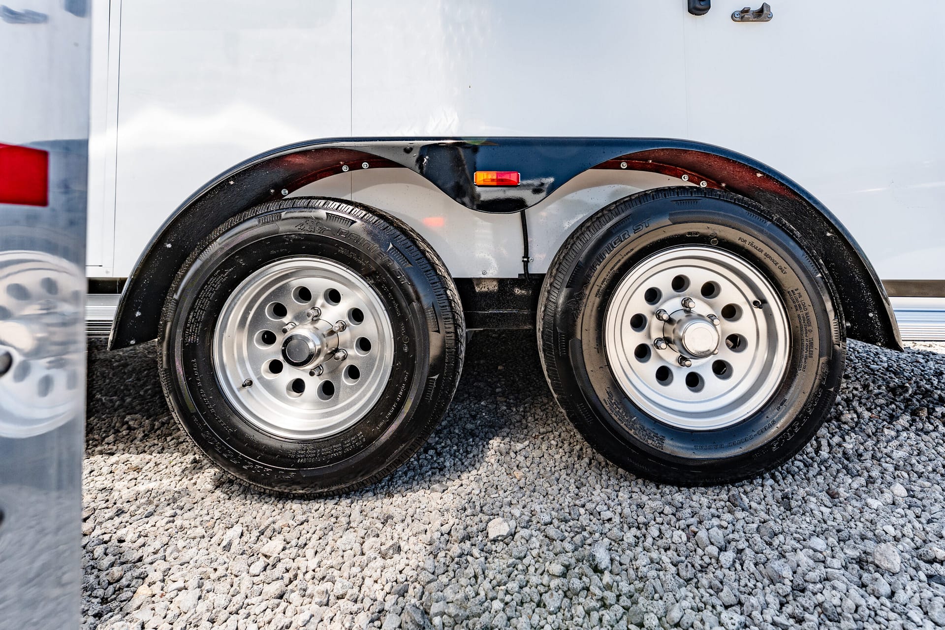 Dual-axle trailer wheels with black fenders on a gravel surface, small orange marker light above the tires.