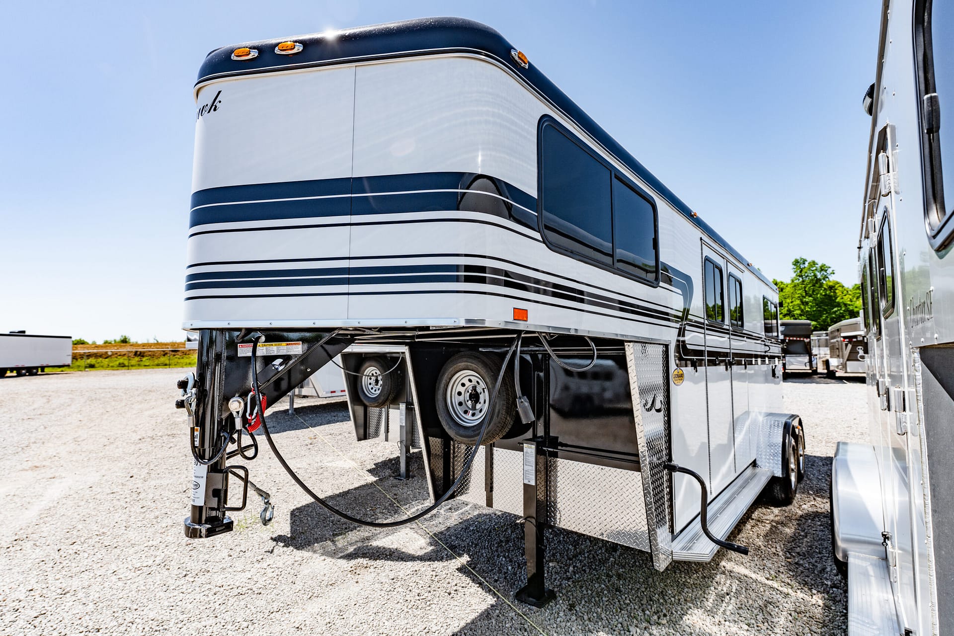 Side view of a large white and blue travel trailer parked on gravel with visible hitch and cables under a clear blue sky.