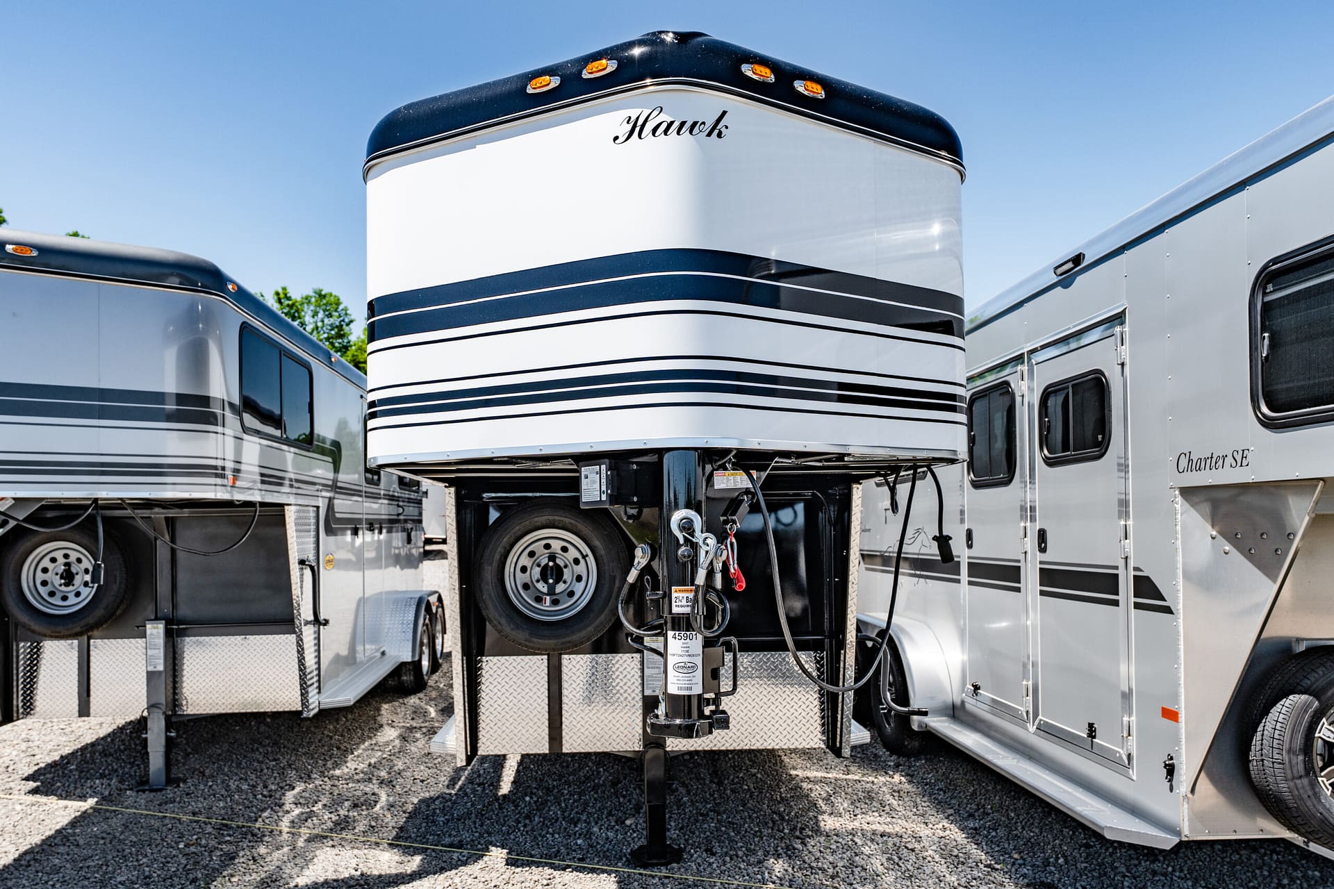 Front view of a Hawk horse trailer with a gooseneck hitch, flanked by silver trailers on a gravel lot.