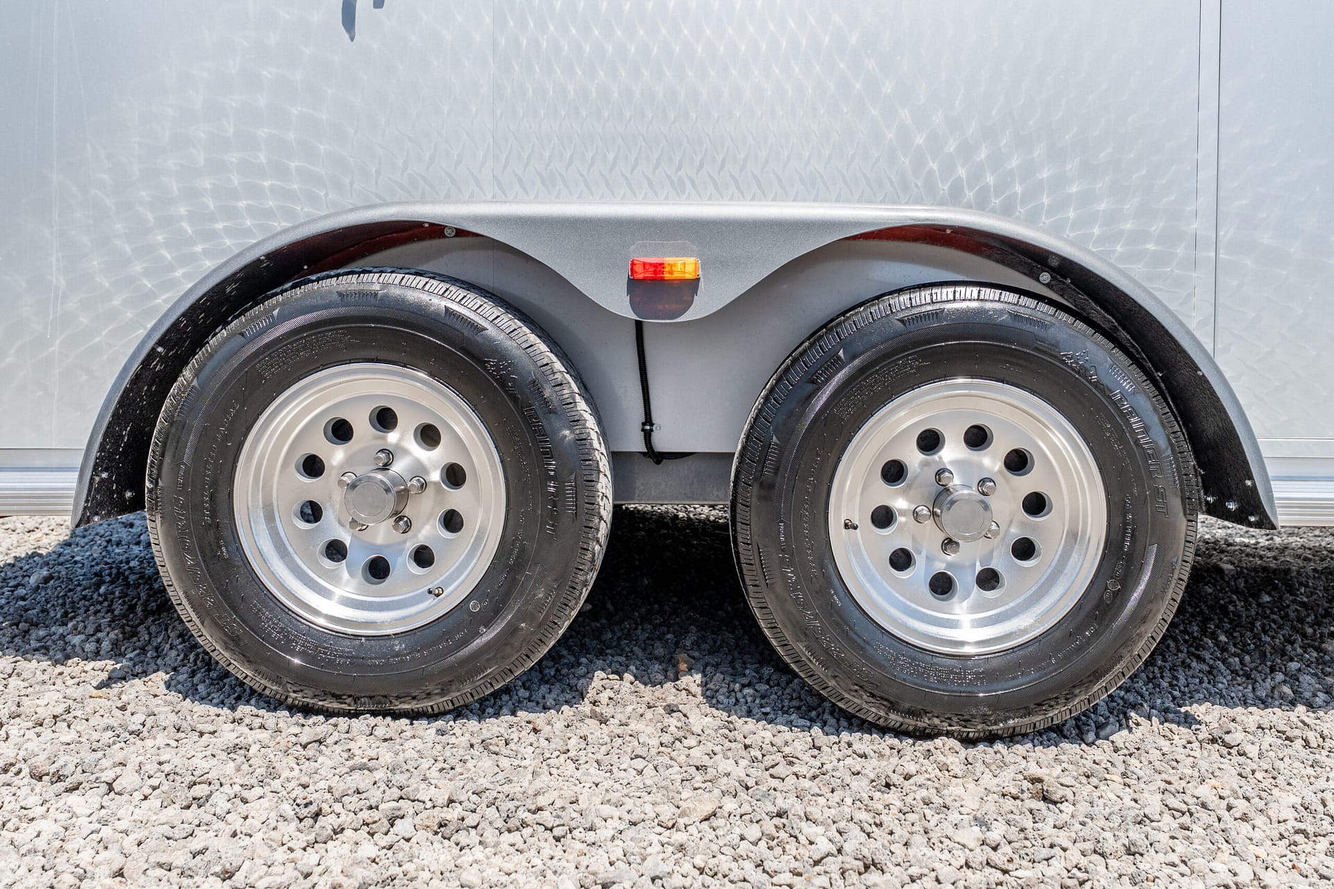 Dual trailer wheels with silver rims under a curved grey fender, small orange marker light centered between tires on a gravel ground.