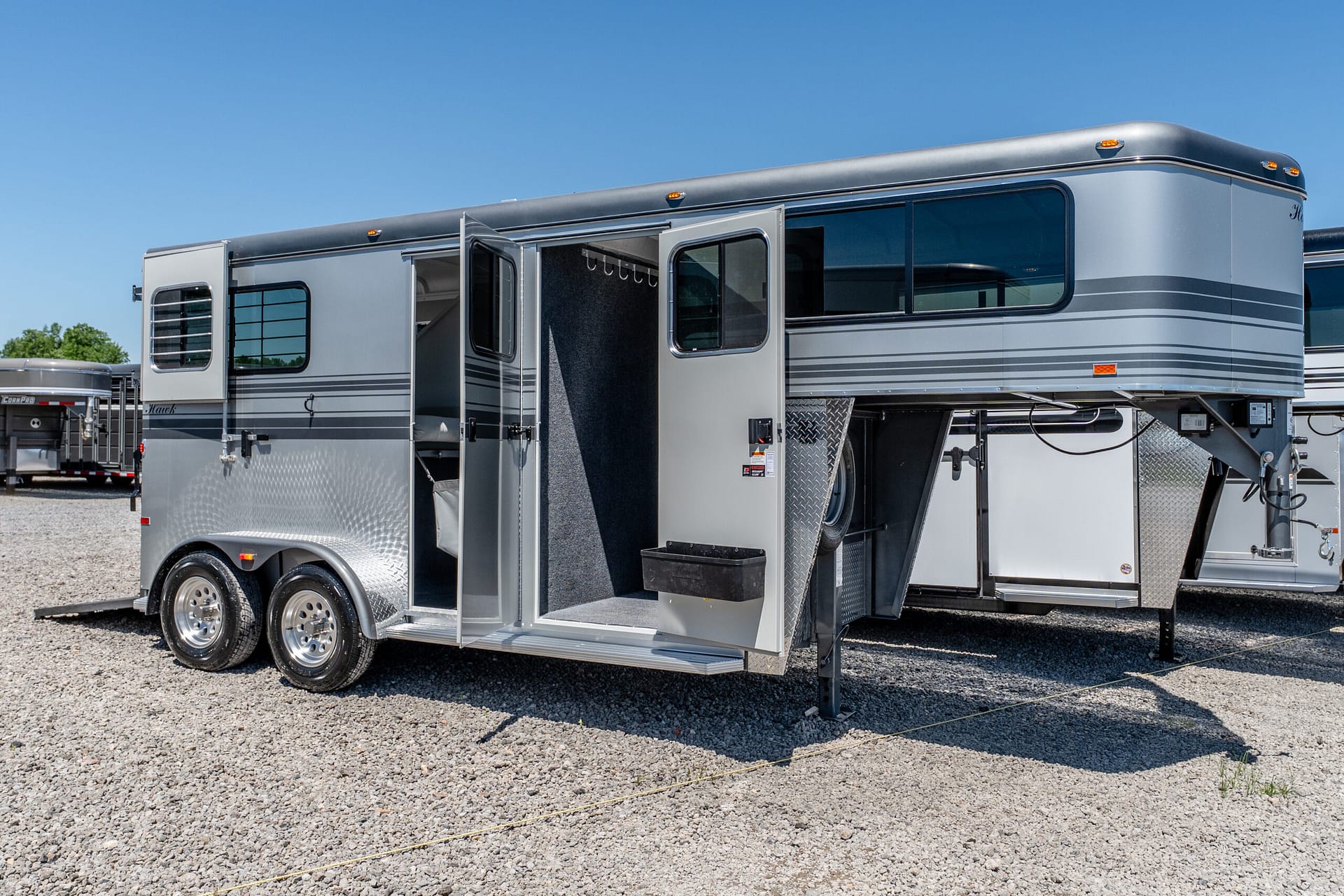 Silver travel trailer with the side door open, exposing interior storage and hooks against a clear blue sky on a gravel lot.
