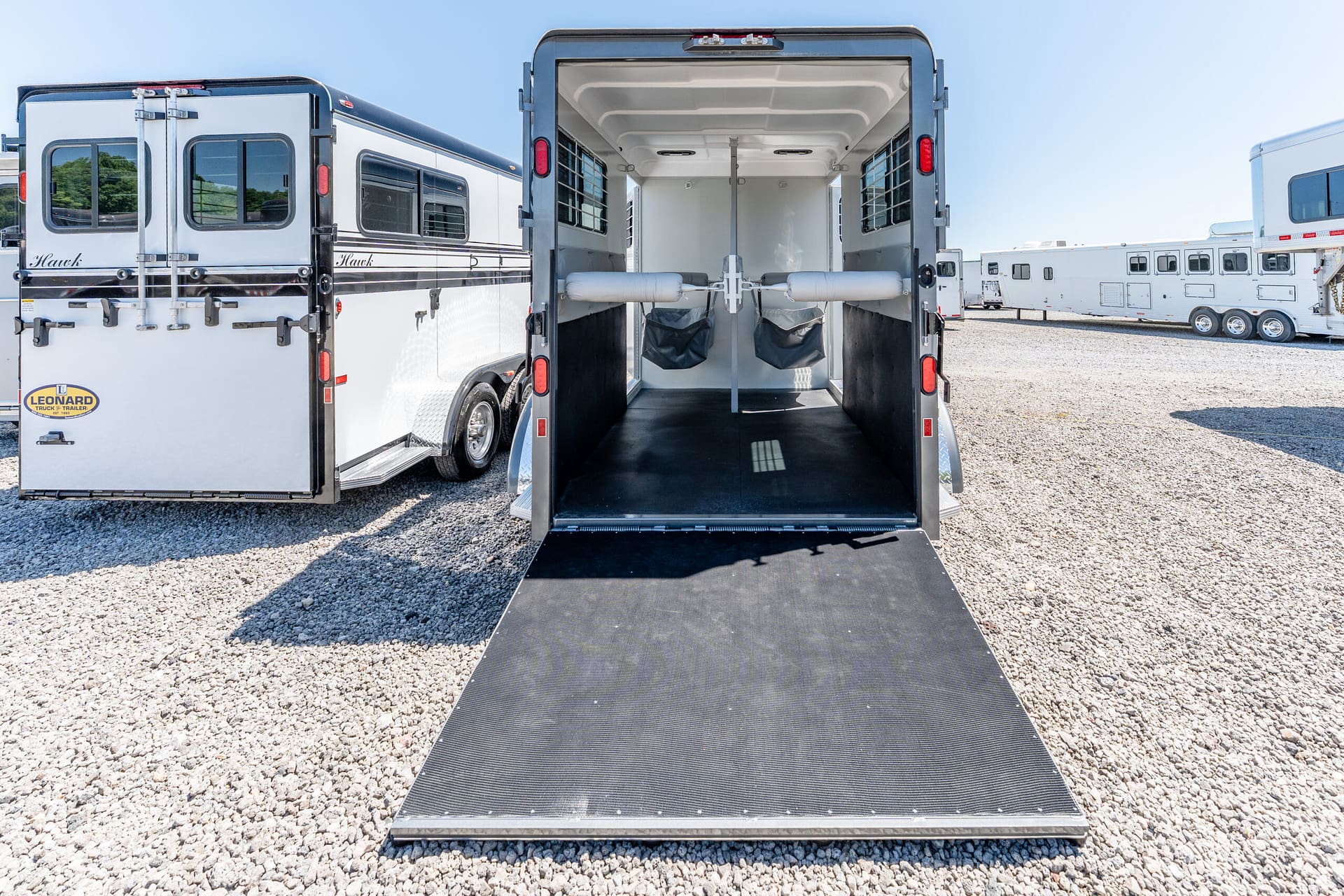 Interior of an open horse trailer showing two padded stalls with center divider and a lowered ramp for loading on gravel lot.