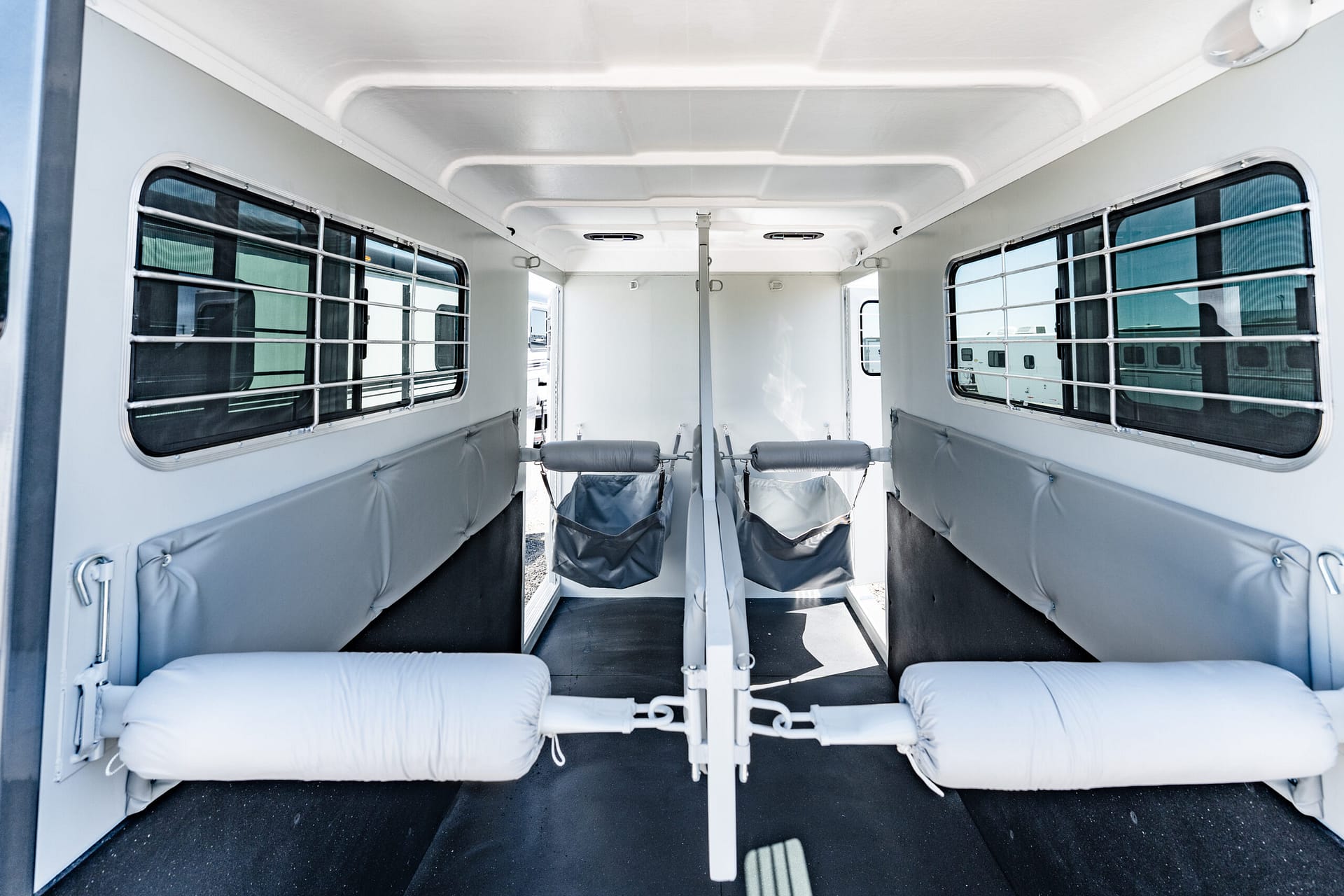 Interior view of a white horse trailer with two padded stalls, metal dividers, and barred windows on both sides; sunlight outside, black non-slip floor.