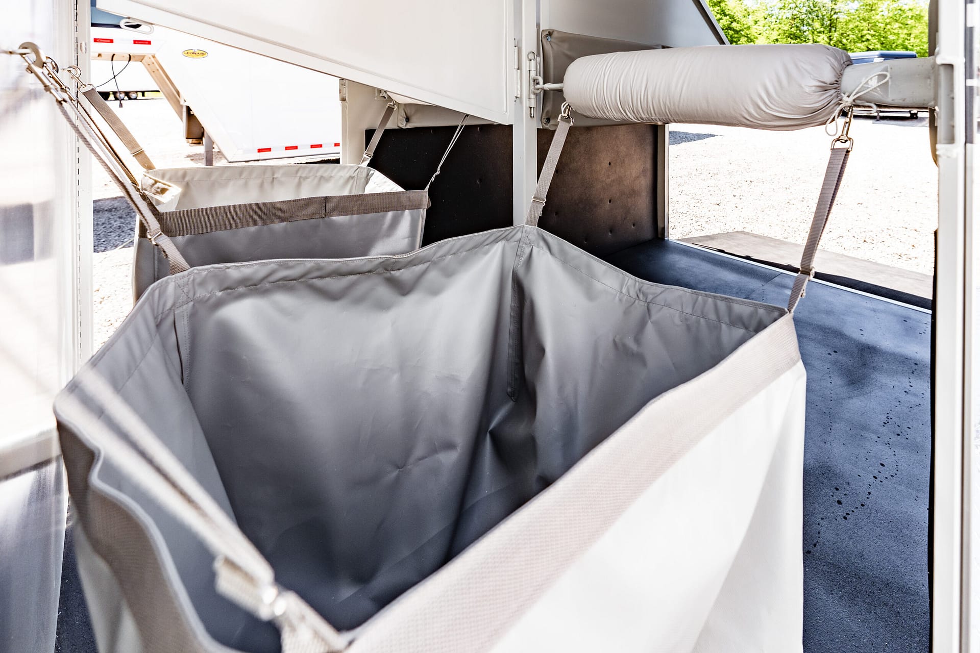 Interior of a trailer showing gray fabric storage bins suspended by adjustable straps along a metal frame, with a padded cushion on a rack and a bright outdoor area visible outside the door.