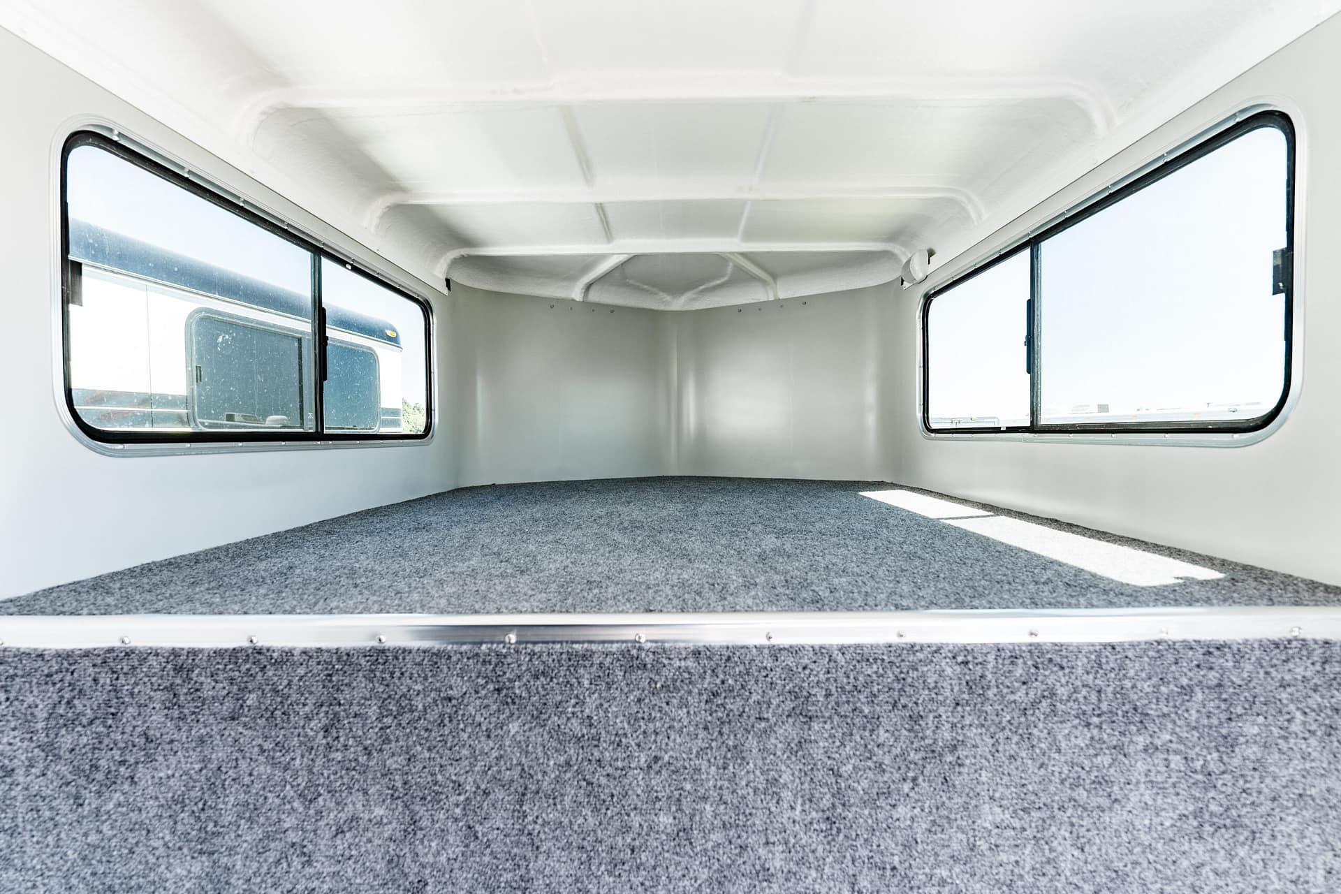 Interior of a small cargo trailer with white walls, two side windows, and a grey carpeted floor.