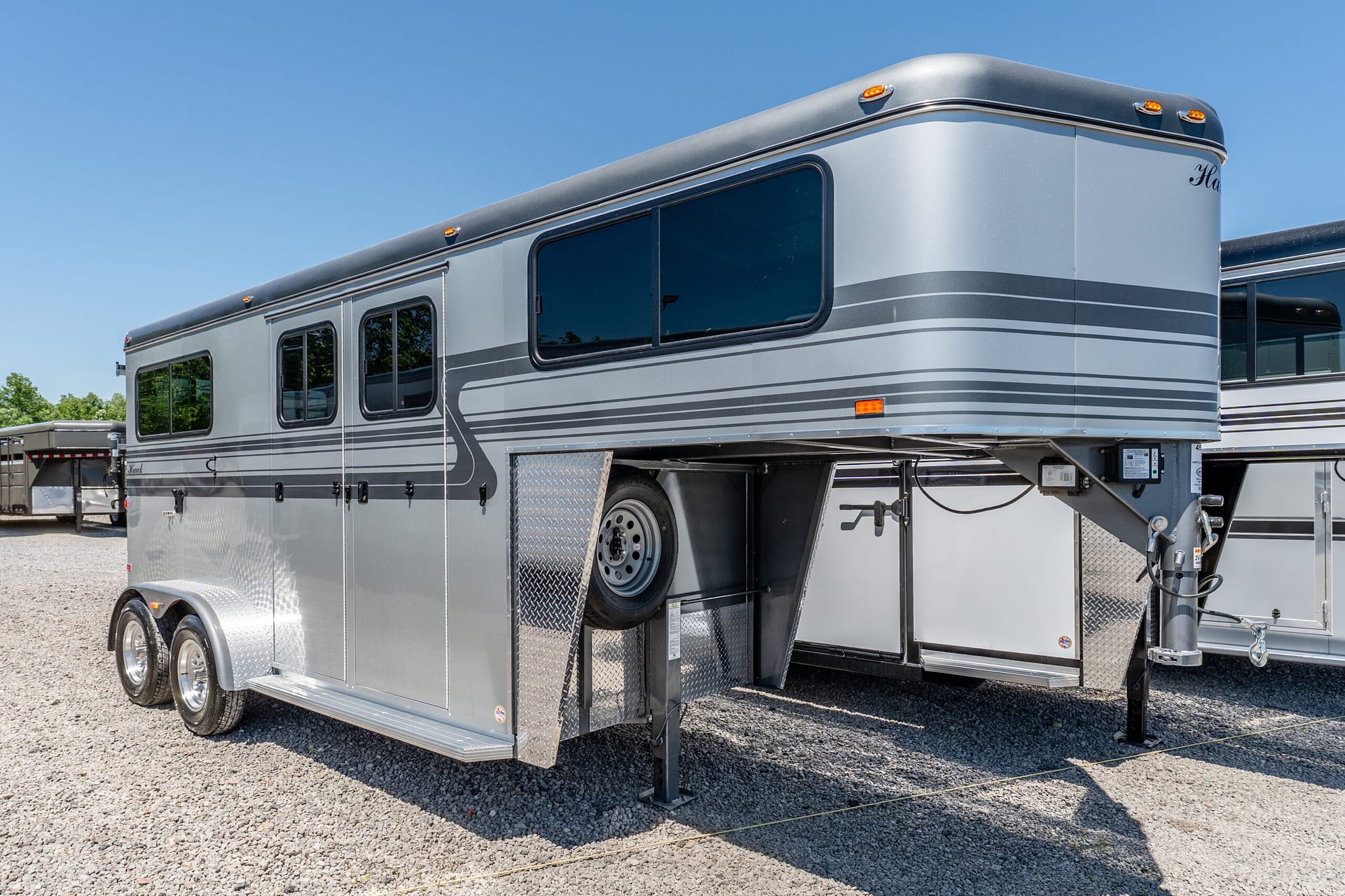 Side view of a large silver gooseneck horse trailer with windows and diamond-plate steps, parked on gravel under a blue sky.
