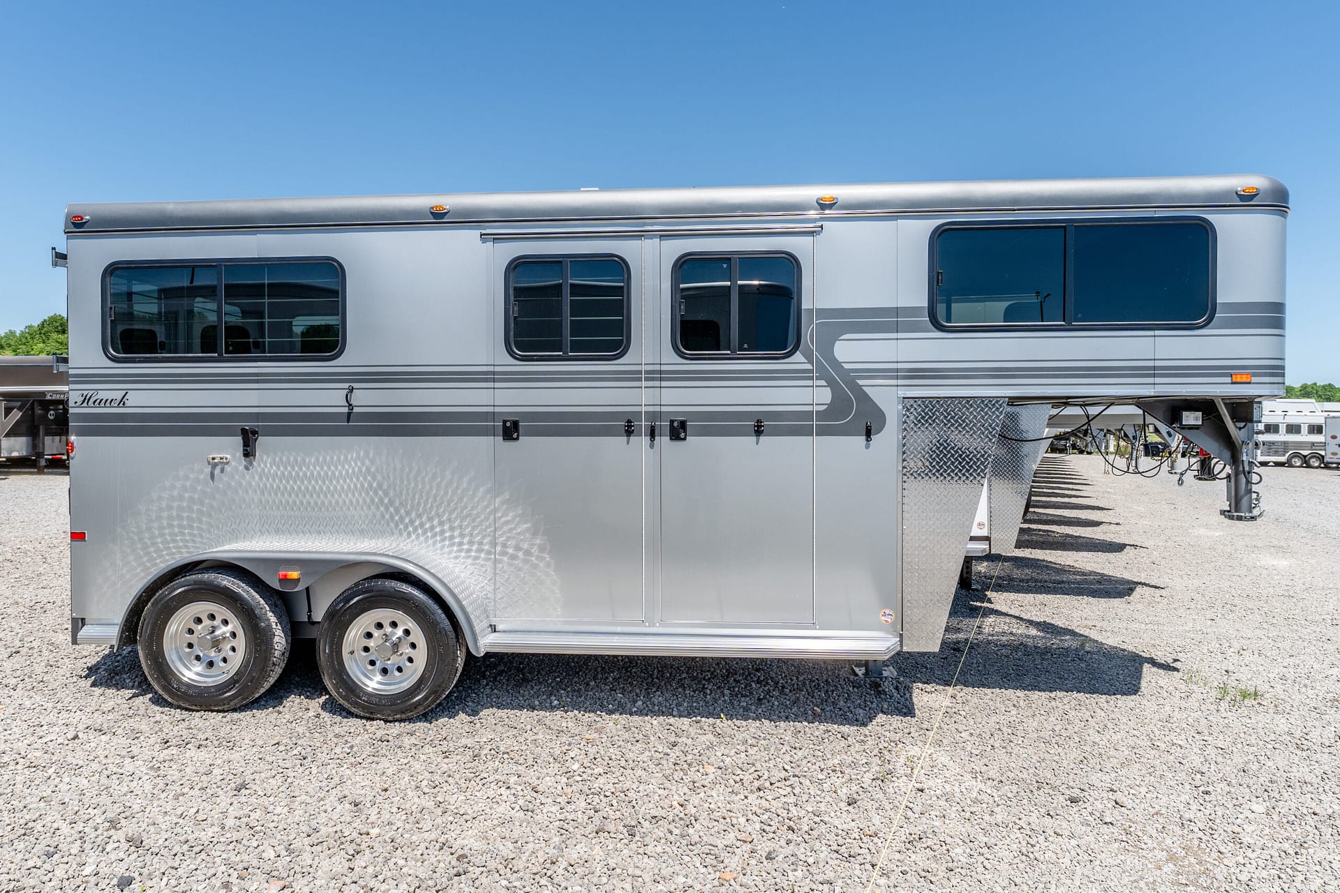 A long silver RV trailer parked on a gravel lot under a clear blue sky, showing windows and wheels on the side.