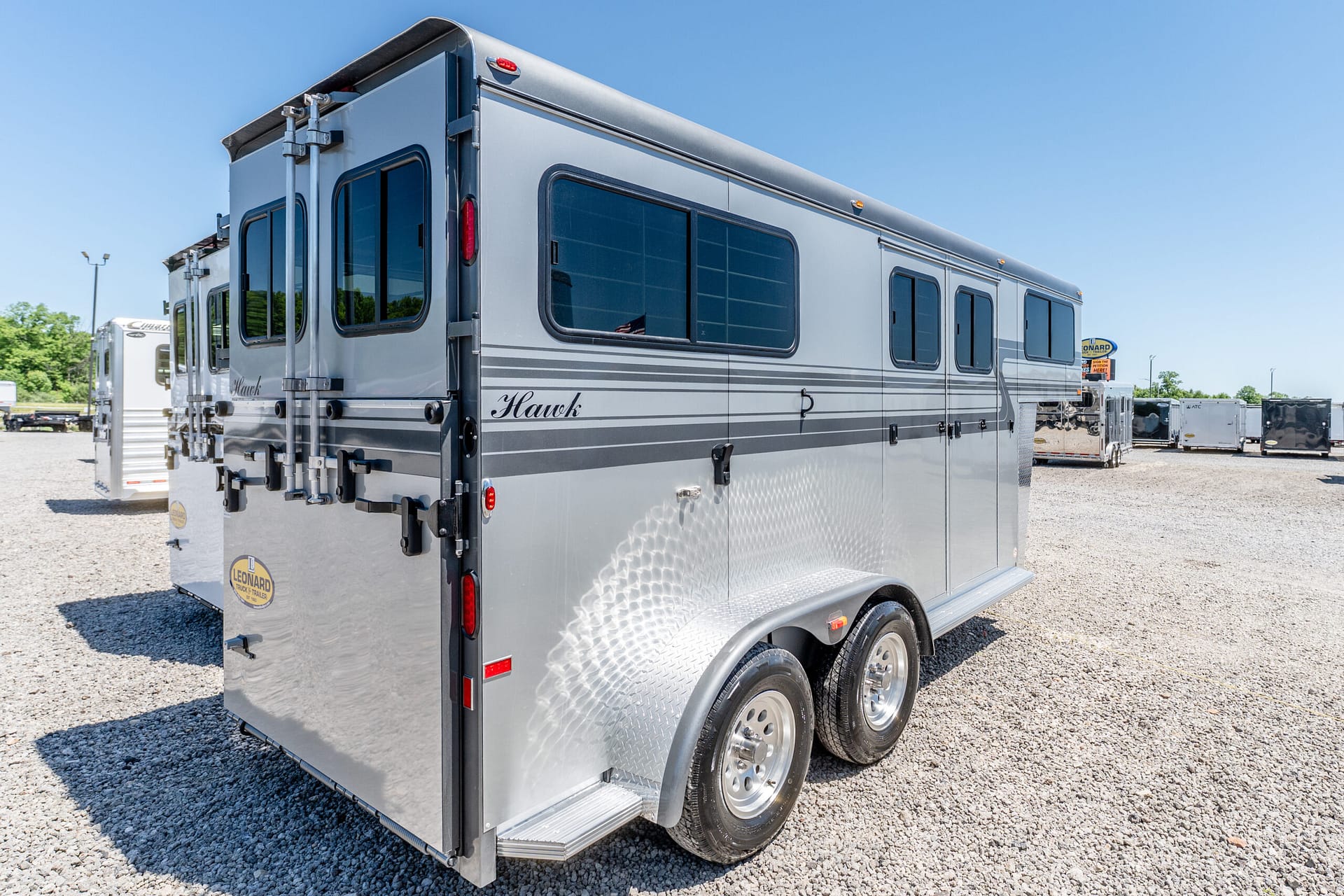 Rear and side view of a silver horse trailer with windows and diamond-plate lower panel parked on a gravel lot under a clear blue sky