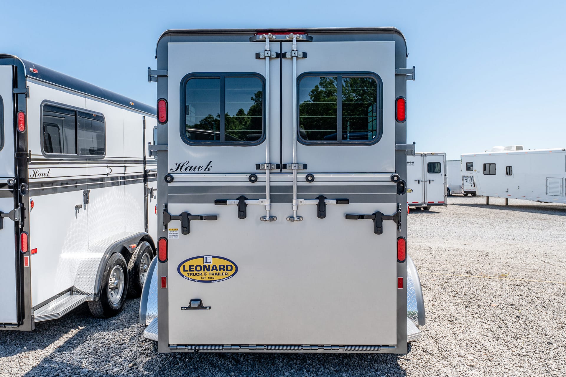 Rear view of a silver trailer with double back doors and two windows, Leonard logo on the lower door, on a gravel lot.