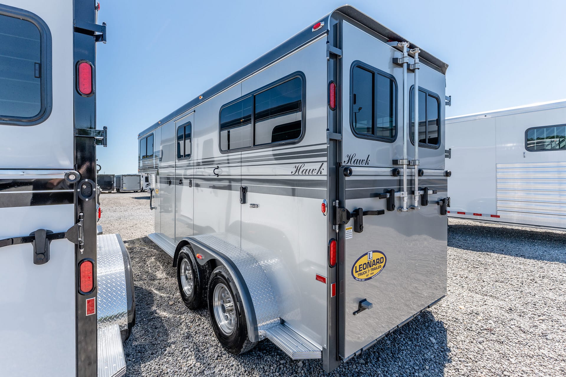 Rear and side of a gray Hawk travel trailer with windows, parked in a lot under a clear sky.